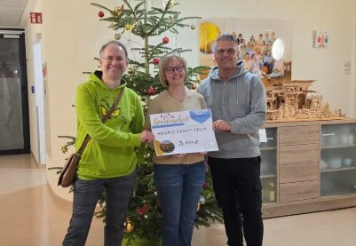 Drei Personen stehen vor einem Weihnachtsbaum, halten einen Scheck und lächeln. Es scheint eine feierliche Übergabe zu sein.