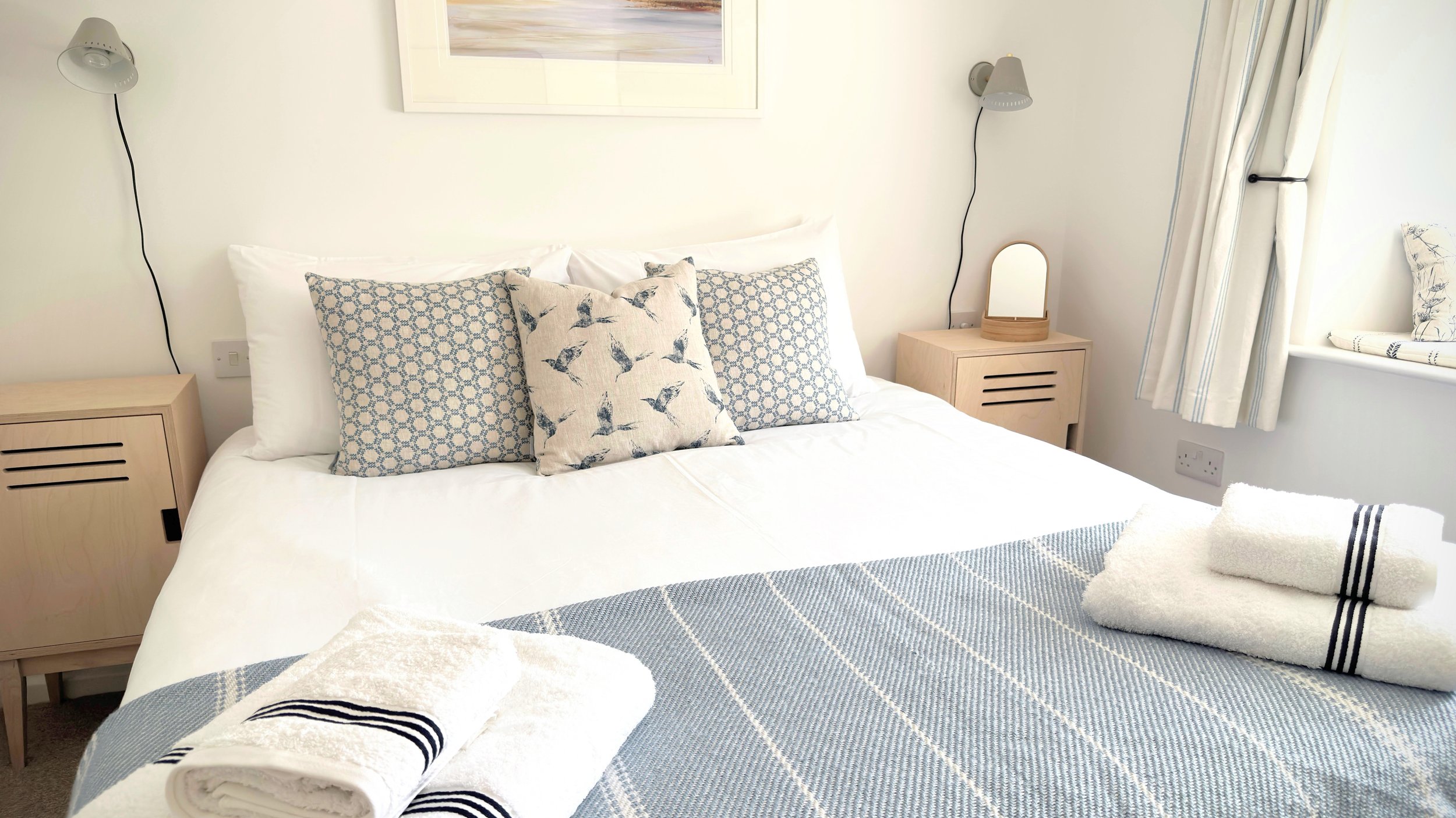 Cozy bedroom with a neatly made bed, featuring white and blue pillows, light wood nightstands, wall-mounted lamps, and folded towels on the bed. Sunlight streams through curtains, creating a serene ambiance.