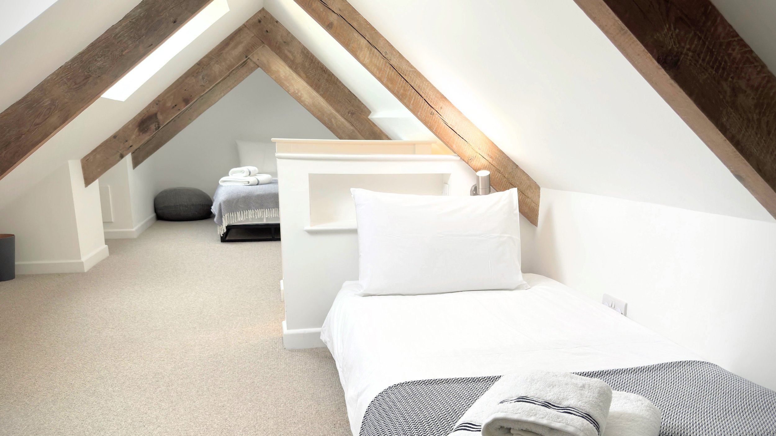 Loft bedroom with wooden beams, single bed with white and gray bedding, and light-colored carpet.