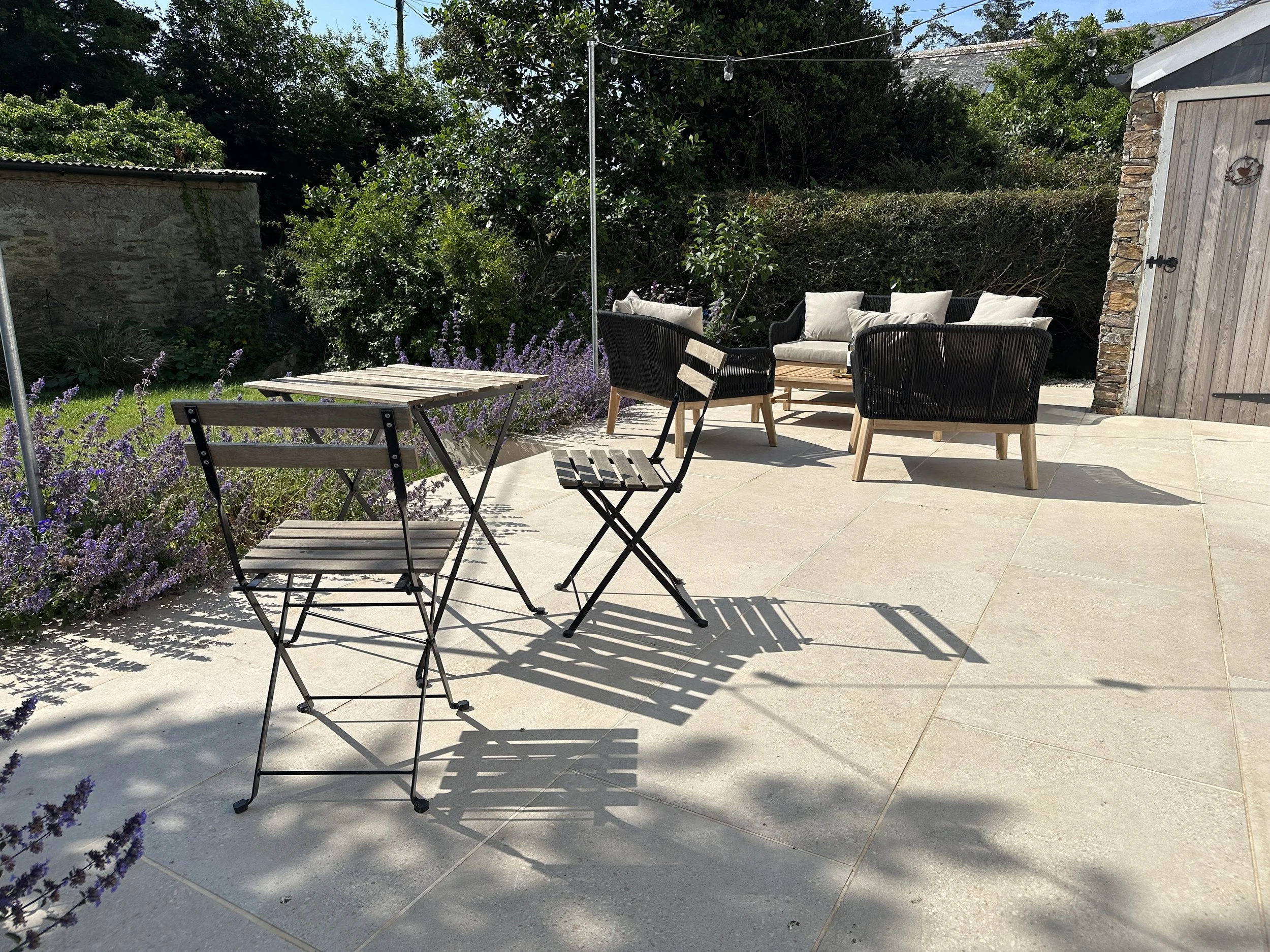 Outdoor patio with a wooden table and chairs, surrounded by lavender plants. Two wicker chairs and a couch with cushions are nearby. A wooden shed is visible in the background.