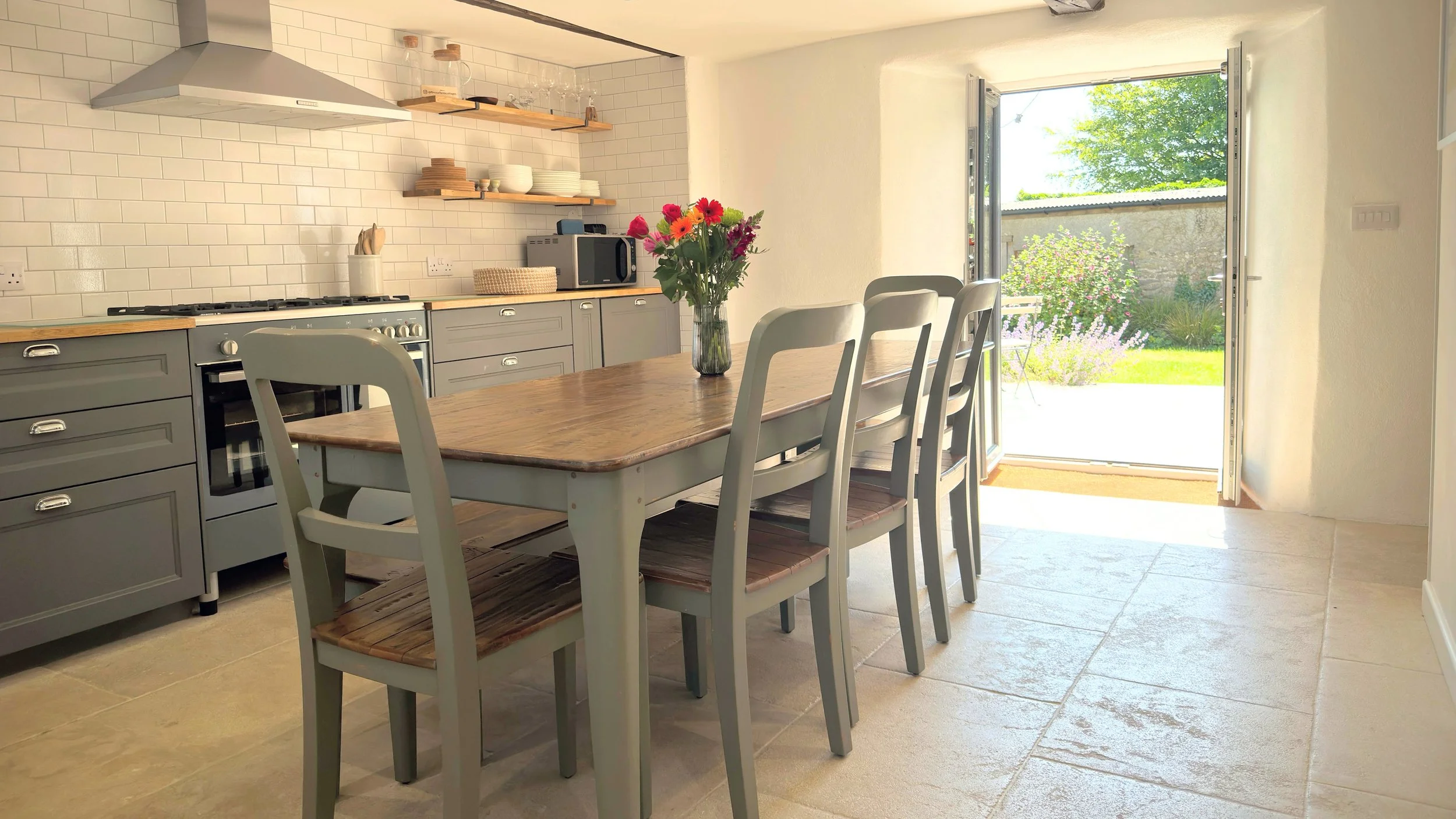 Modern kitchen with grey cabinets, wooden dining table set, open glass doors leading to a garden, and a vase of flowers on the table.