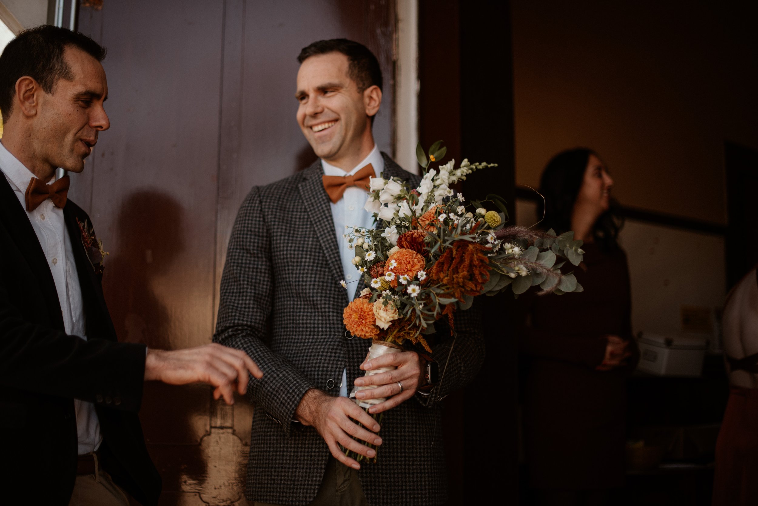 Deux hommes en costumes avec des nœuds papillon bruns, un tenant un bouquet de fleurs orange et blanches, souriant dans une ambiance chaleureuse.