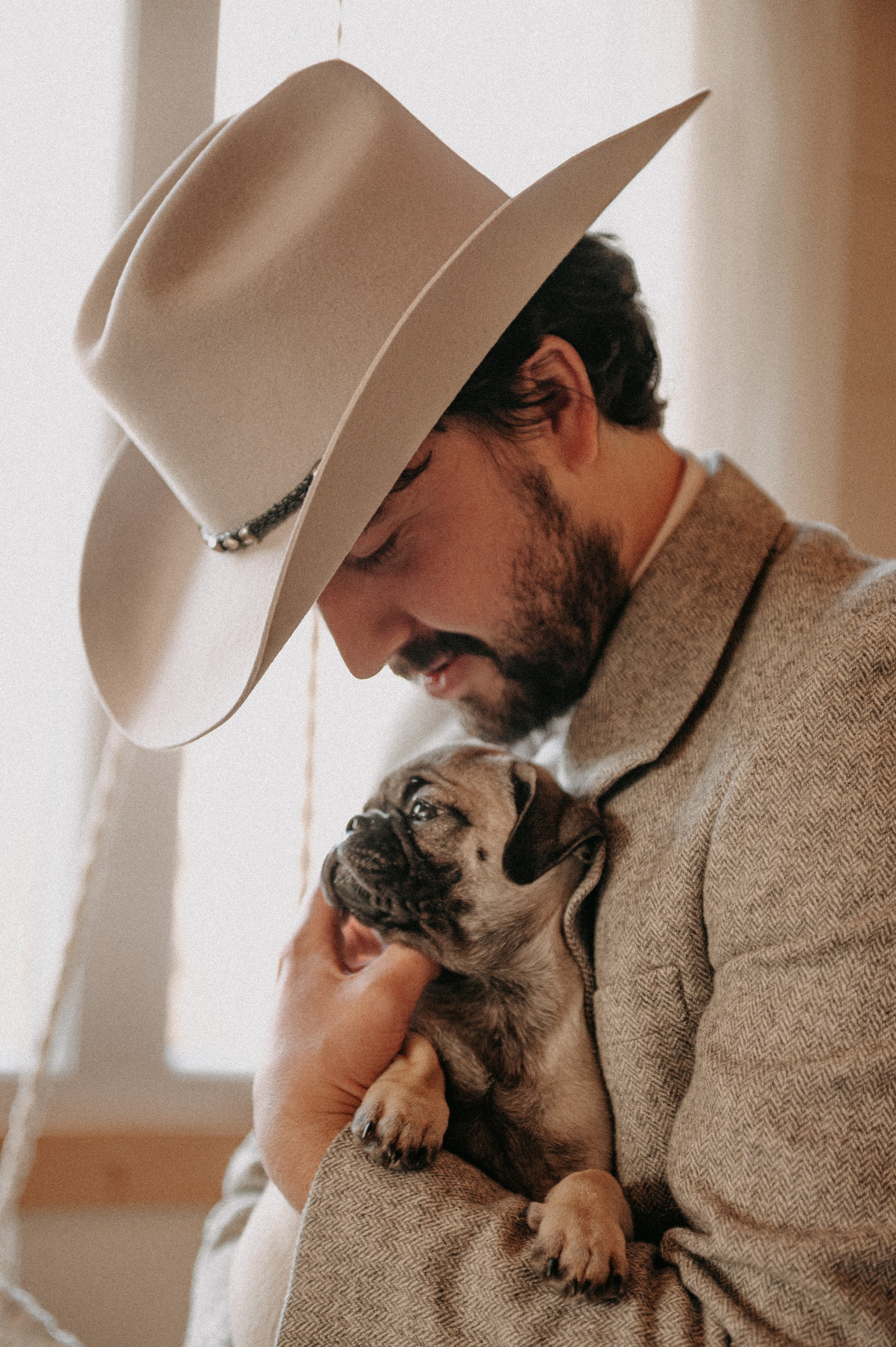 Un homme portant un chapeau beige tient un petit chiot mignon dans ses bras, à l'intérieur d'une maison.