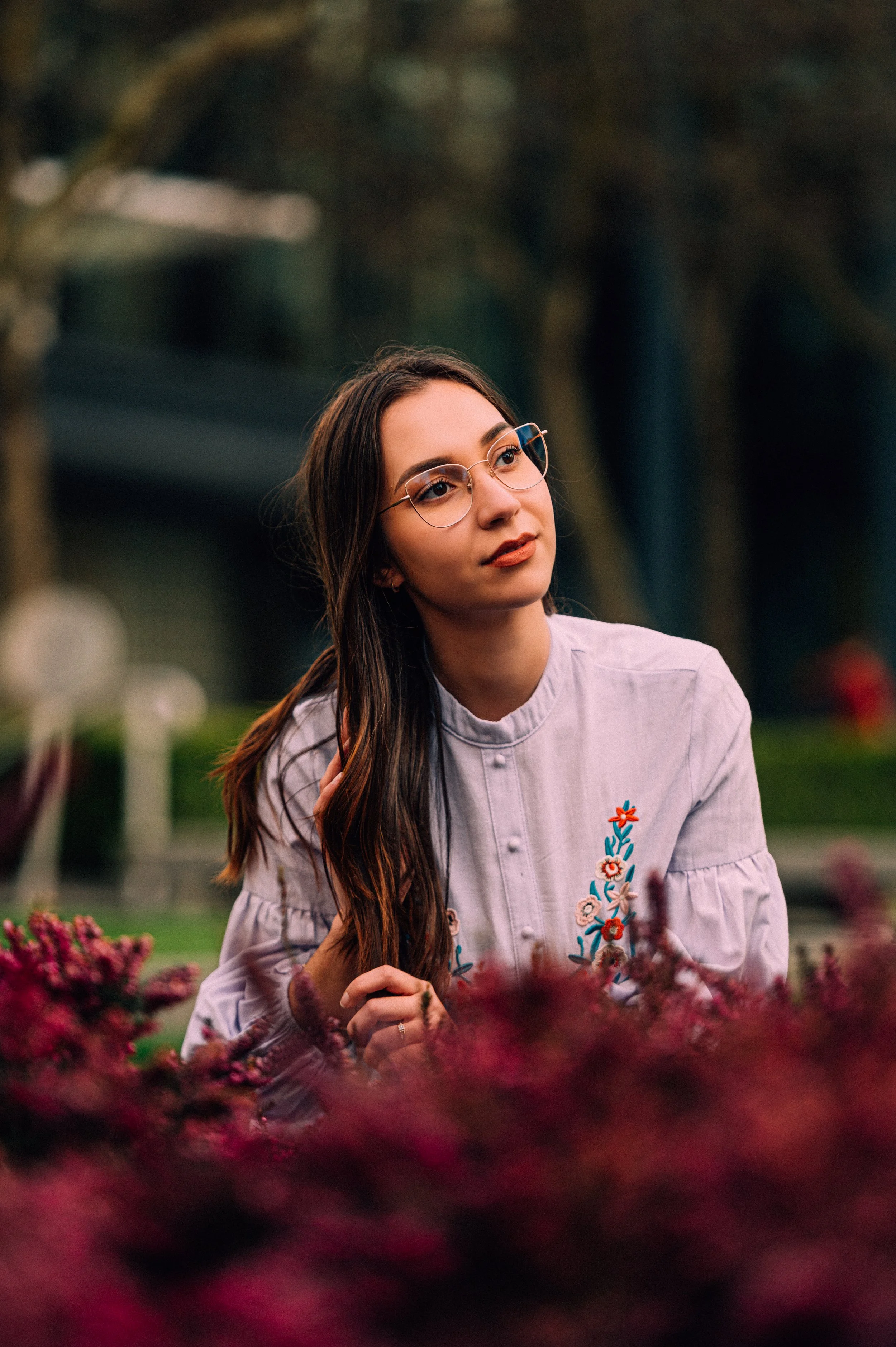 Jeune femme aux lunettes assise parmi des fleurs violettes dans un parc, regardant pensivement au loin.