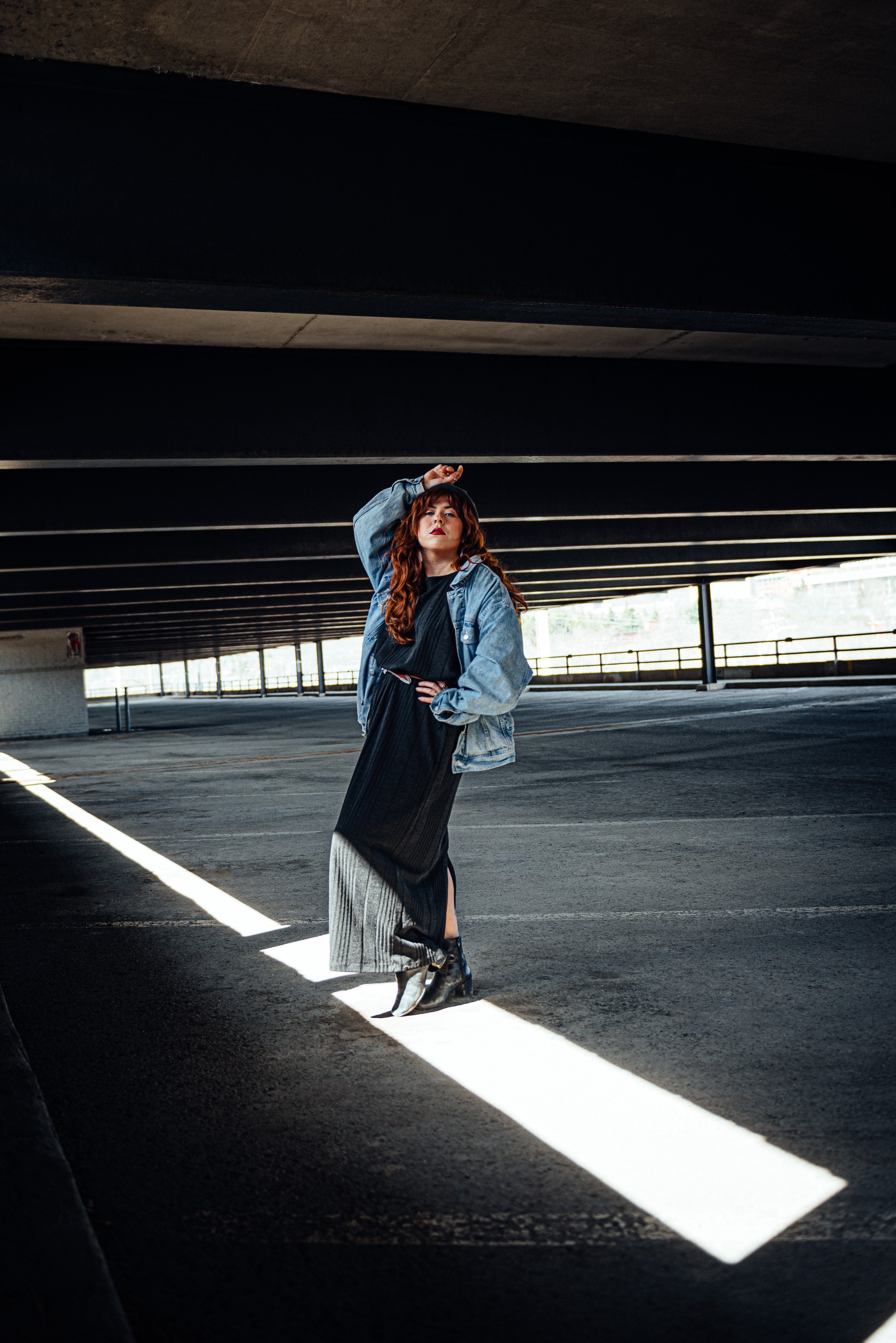 Portrait mode d'une femme aux cheveux roux portant une veste en jean, une robe noire et des bottes noires, se tient dans un parking sous un bâtiment, avec une lumière naturelle derrière elle.