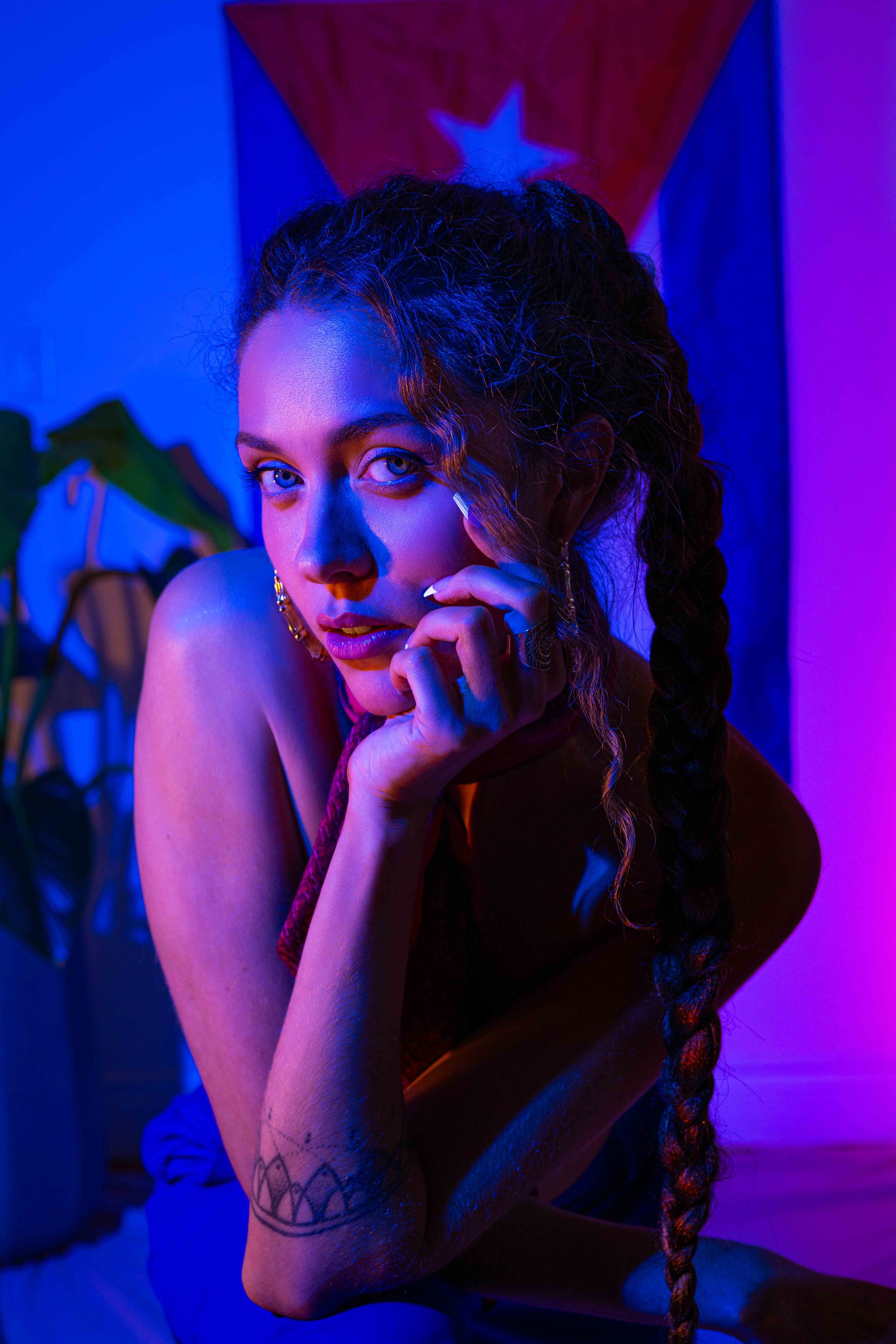 Jordana D'Amours, professeure de danse et danseuse aux cheveux bouclés, portant un haut noir et une longue jupe rouge, assise près d'une fenêtre avec des rideaux, regardant vers la gauche.