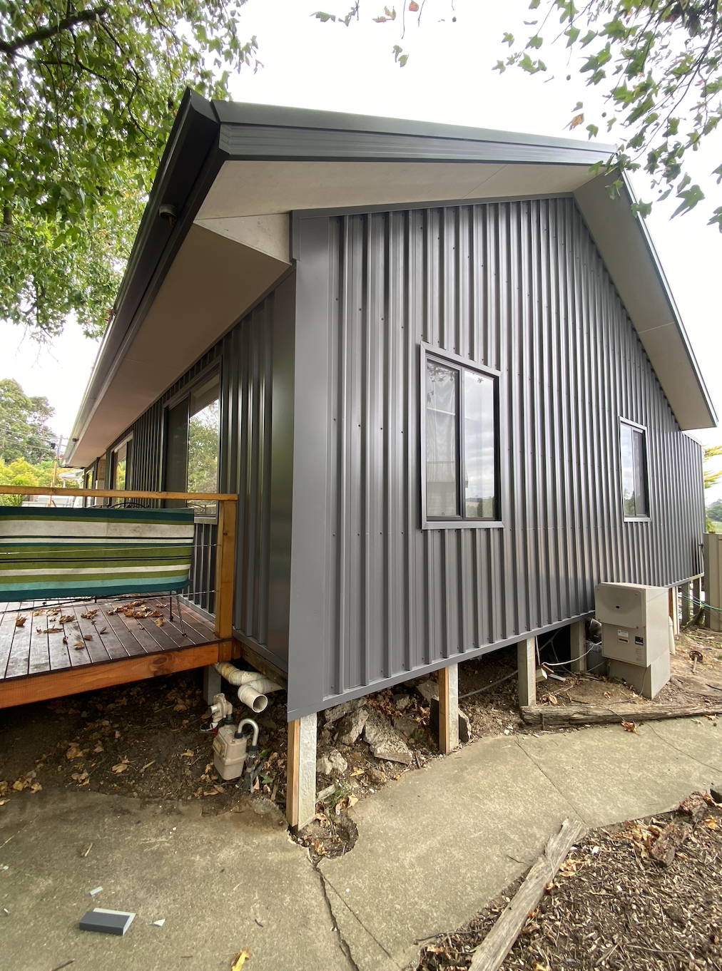 Modern gray metal-clad house with a small wooden deck, surrounded by trees and a concrete path.