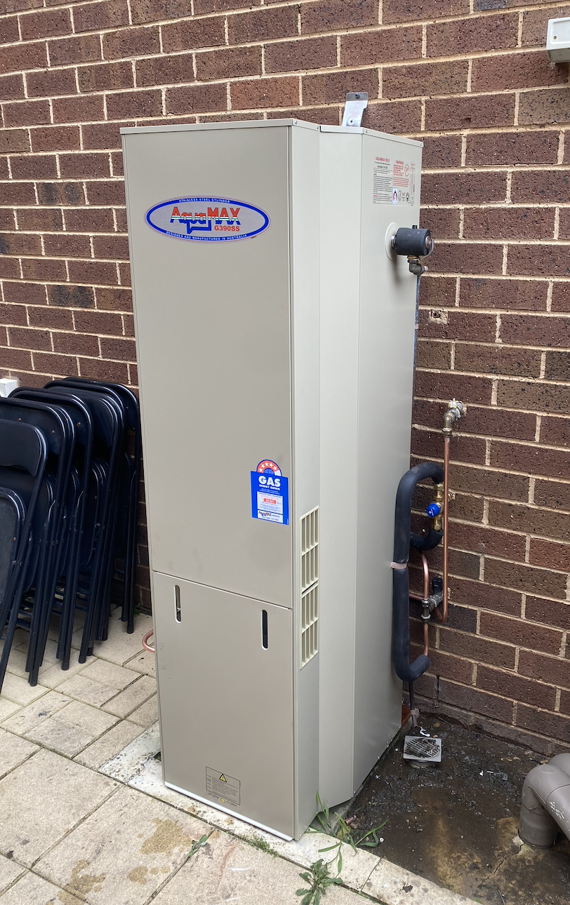Outdoor gas hot water system installed next to a brick wall, with pipes and valves visible, and stacked chairs nearby on a paved surface.