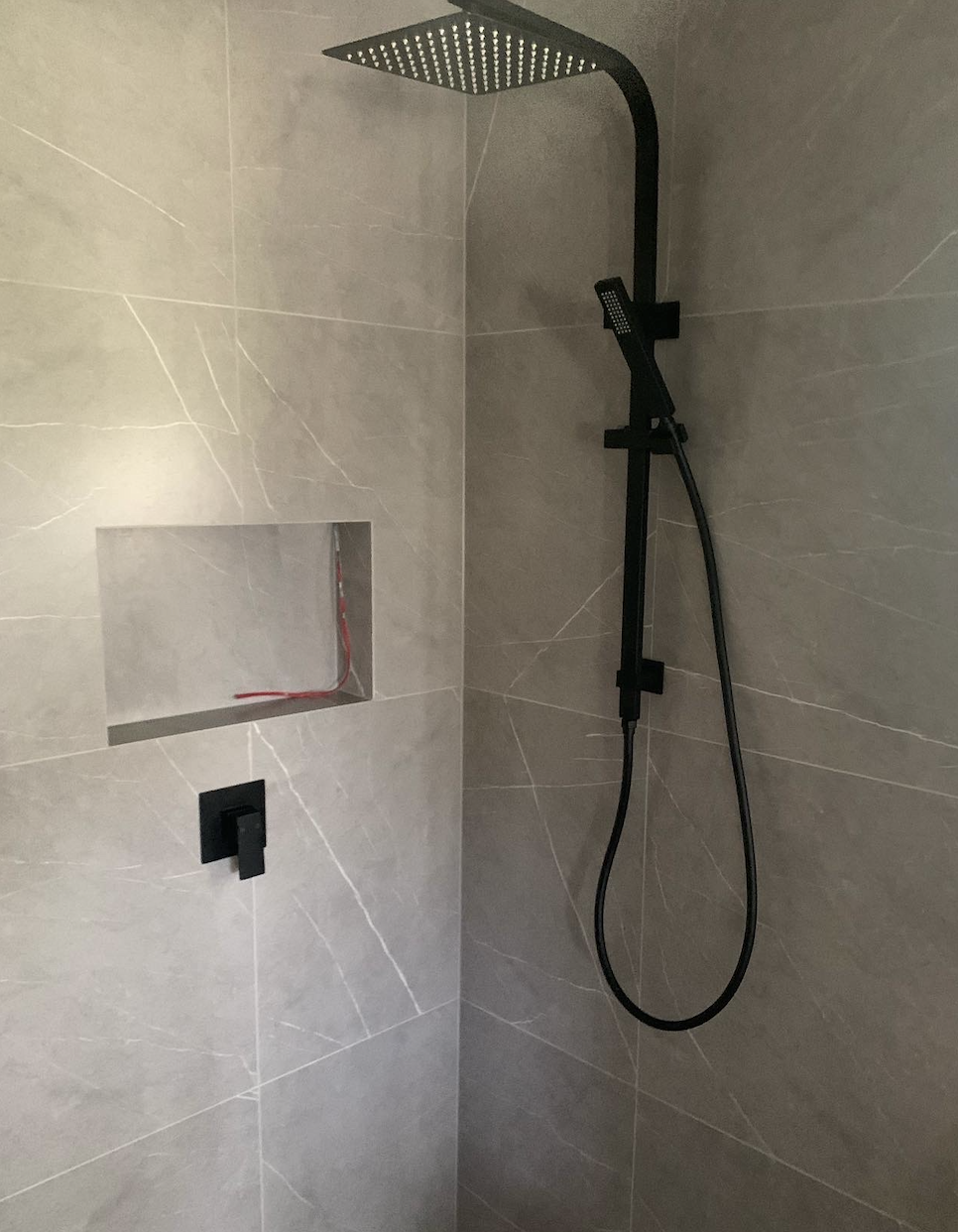 Modern shower with black square rainfall showerhead and handheld attachment, gray tiled walls, and built-in niche.