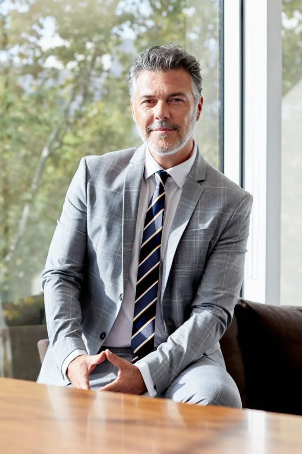 A middle-aged man in a gray plaid suit with a striped tie, sitting on a chair in front of a large window with trees outside, looking at the camera.