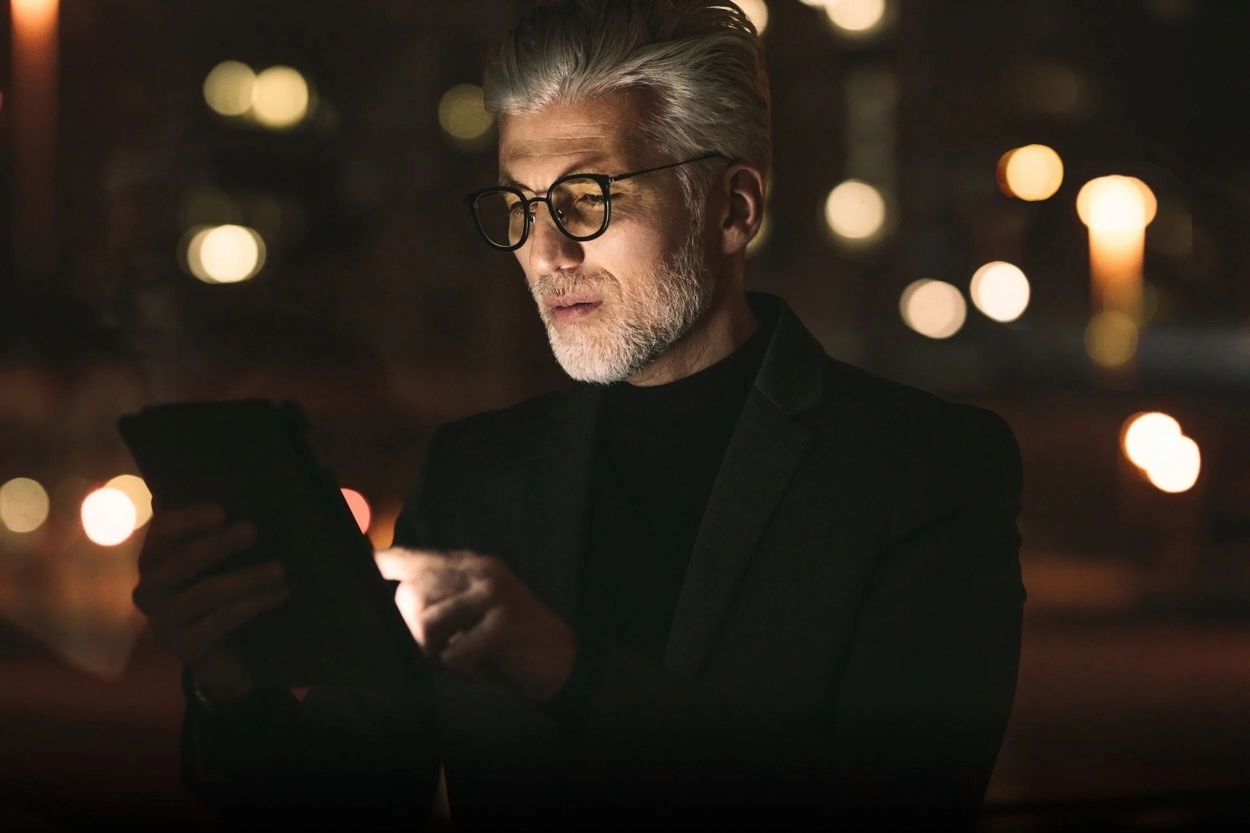 An older man with grey hair and beard, wearing glasses and a black blazer, stands outdoors at night, looking at his phone with blurred city lights in the background.