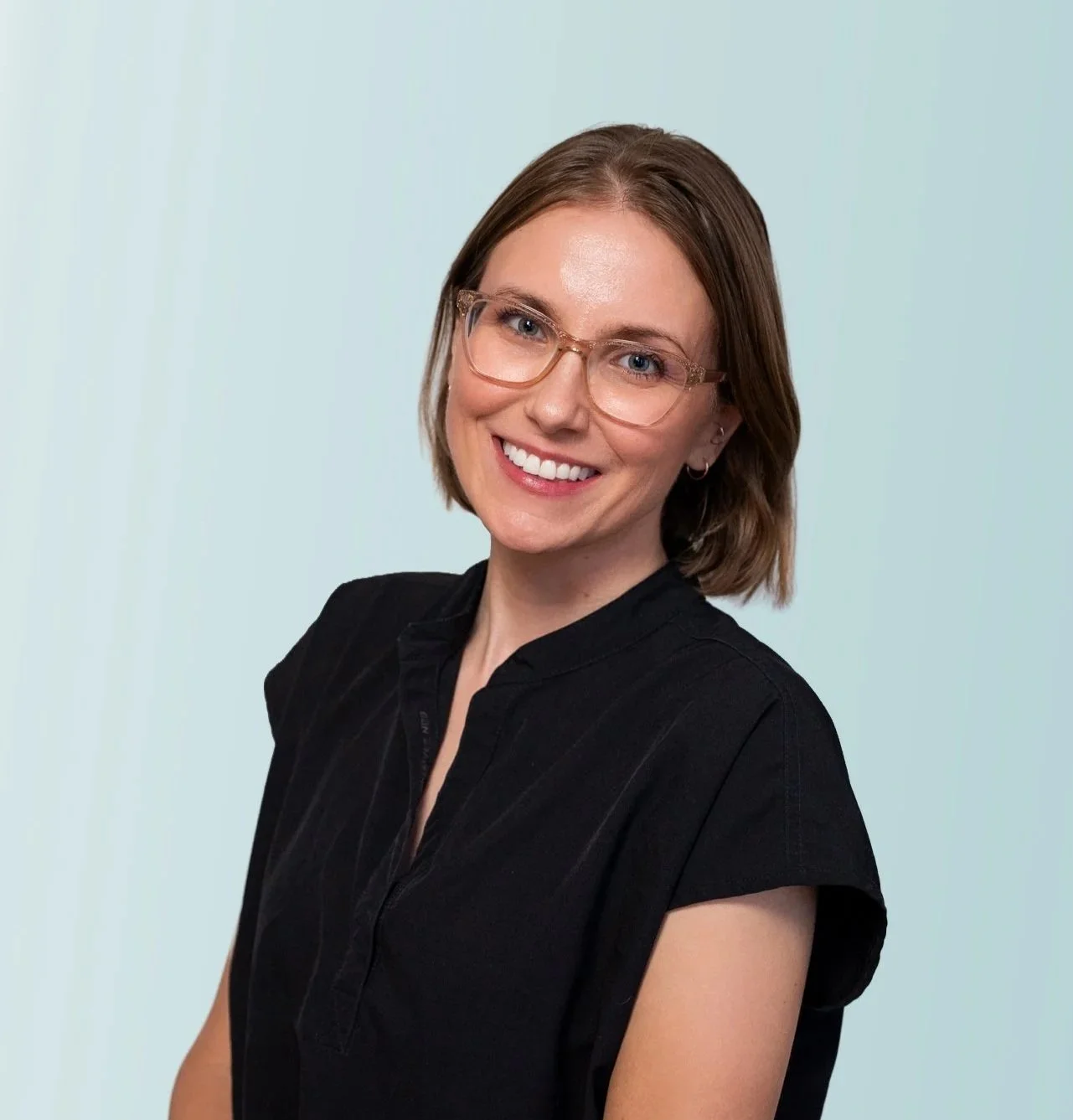 A young woman with shoulder-length hair, wearing glasses and a black top, sitting on a wicker chair against a light blue background, smiling at the camera.
