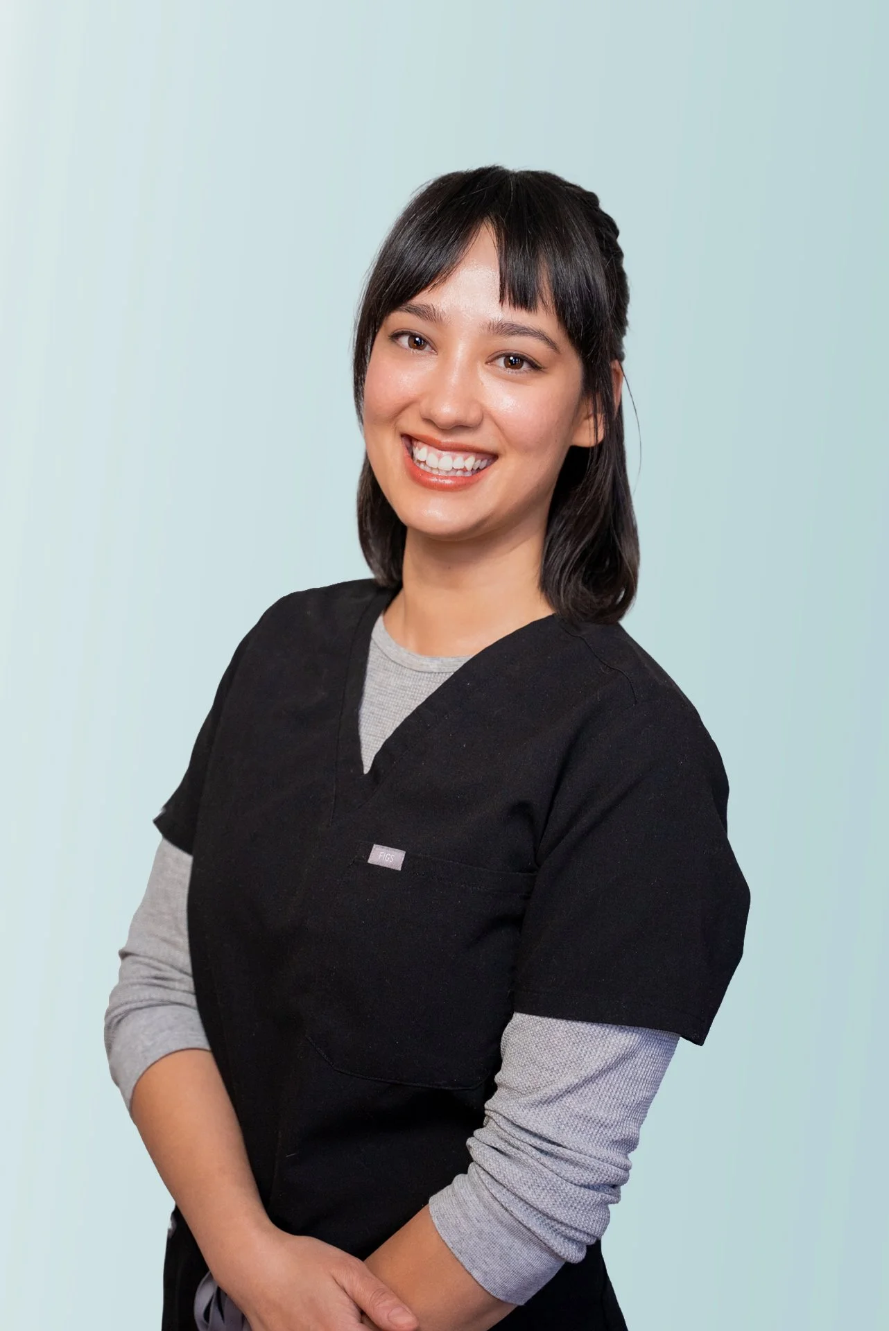 Photo of acupuncturist smiling with long dark hair. Her arms are crossed, she is wearing black scrubs. 