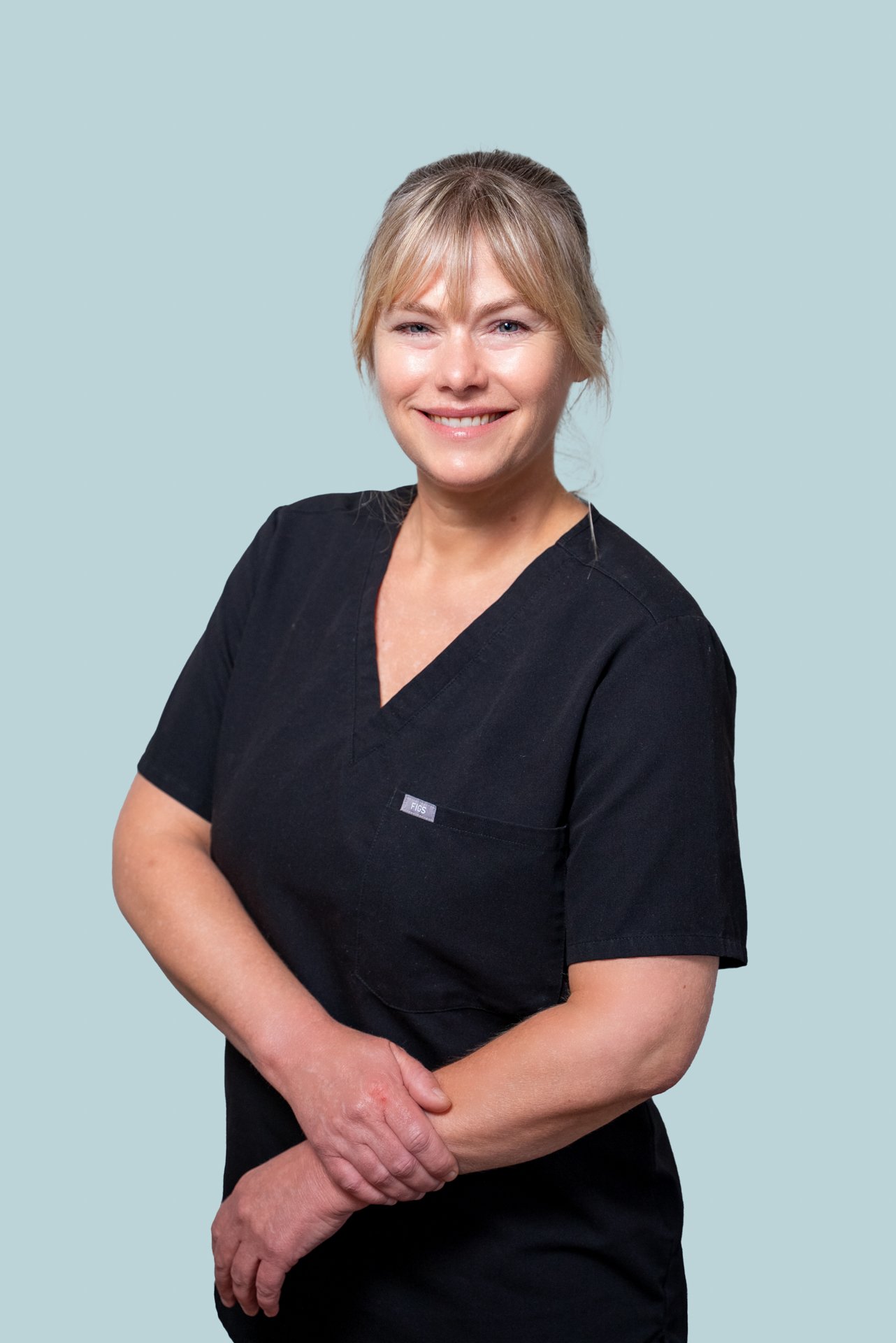 A smiling woman with blonde hair wearing a black work uniform stands with her arms crossed against a light blue background.