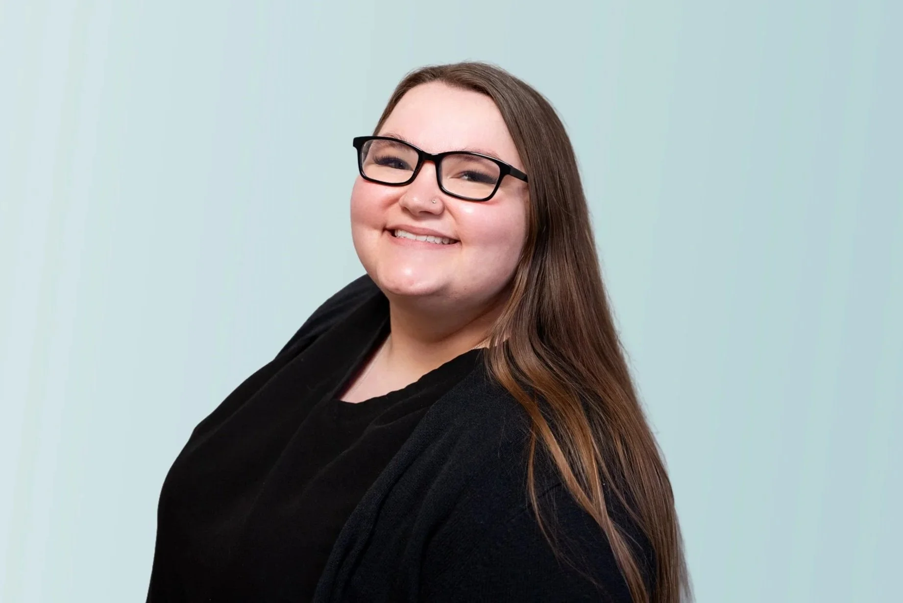 A smiling woman with long brown hair, wearing black glasses, a black top, a necklace with a heart-shaped pendant, and makeup including false eyelashes, standing against a plain beige wall.