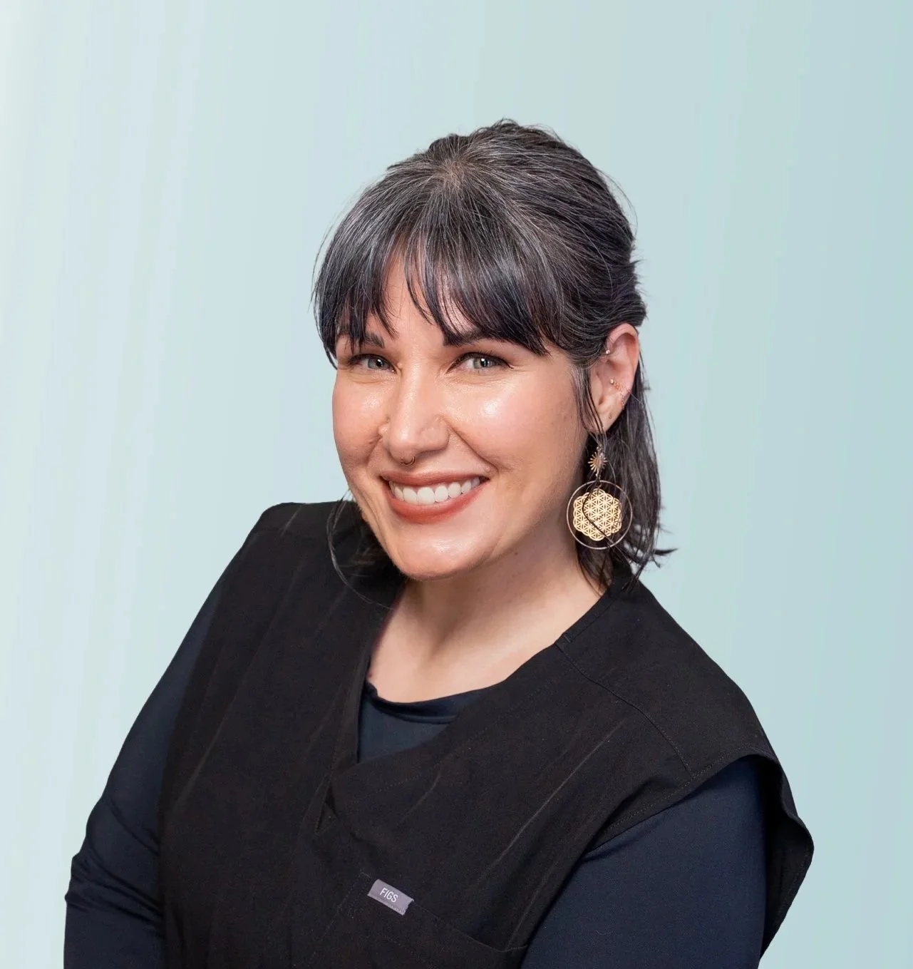 Smiling woman with shoulder-length dark hair featuring purple streaks, wearing a black jacket and a green polka dot skirt, standing against a light blue background.