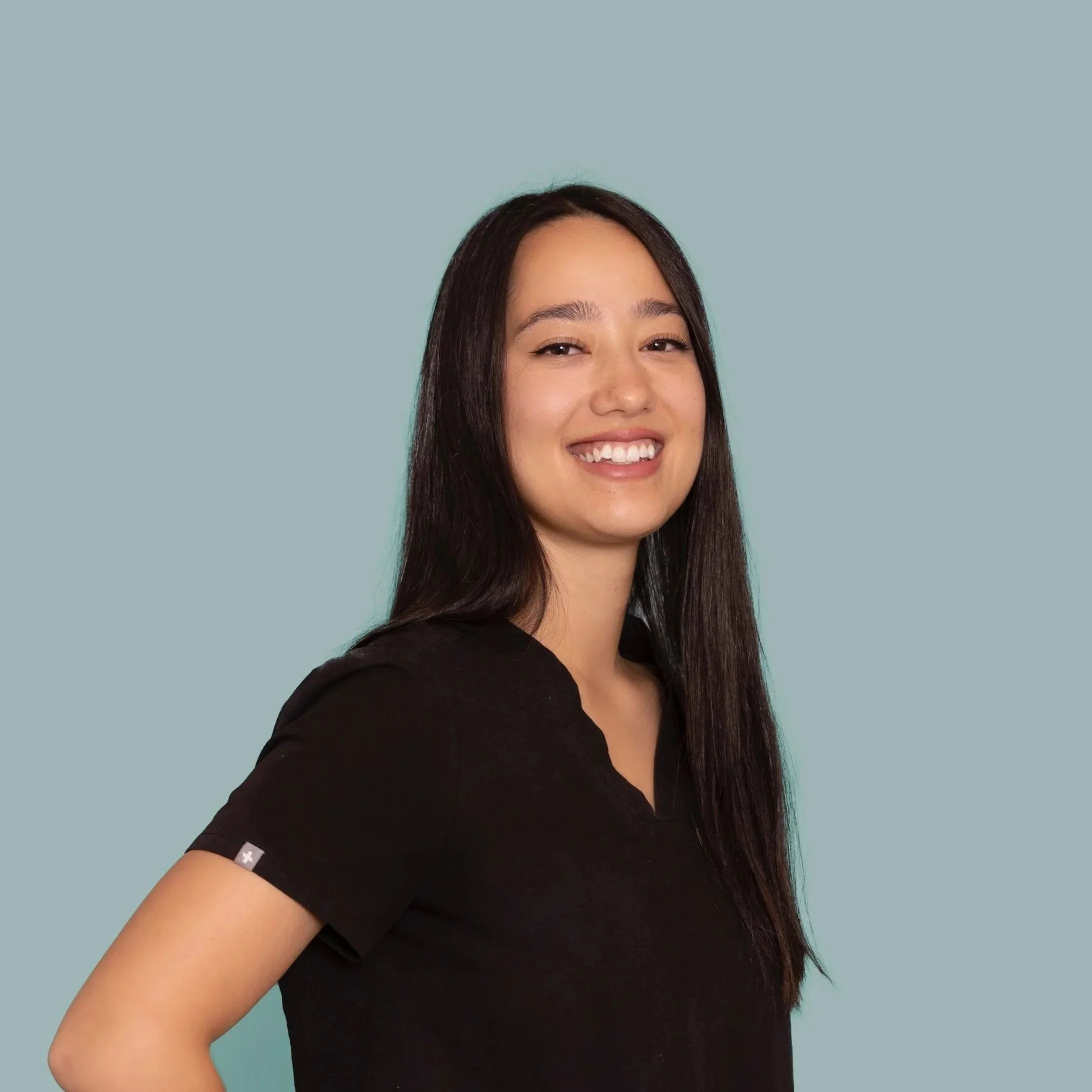 Photo of acupuncturist smiling with long dark hair. Her arms are crossed, she is wearing black scrubs. 