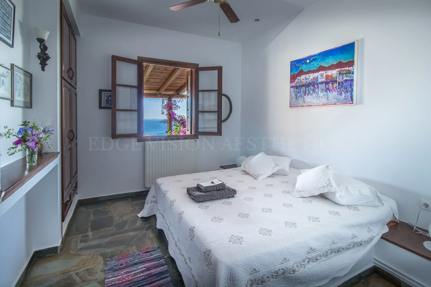A cozy bedroom with white walls, a bed covered in white patterned bedding, and pillows. An open window shows a view of the ocean, wooden roof, and pink bougainvillea outside. There is a colorful landscape painting on the wall, a small side table, and