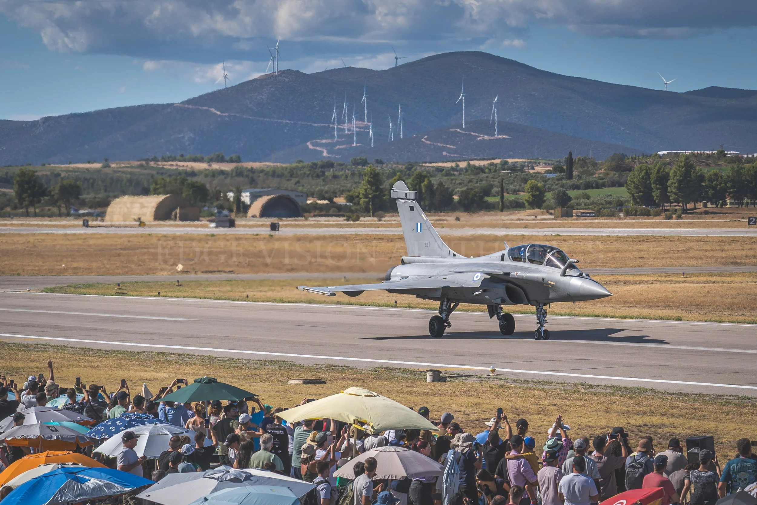 Crowd gathered at an airshow watching a fighter jet on the runway with mountains and wind turbines in the background.