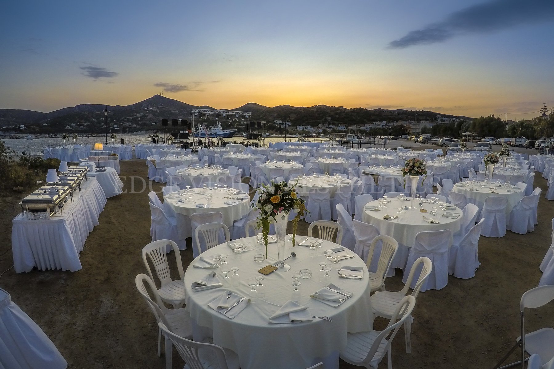Setup of round tables with white tablecloths, chairs, and floral centerpieces on a sandy outdoor area overlooking water, mountains, and sunset sky.