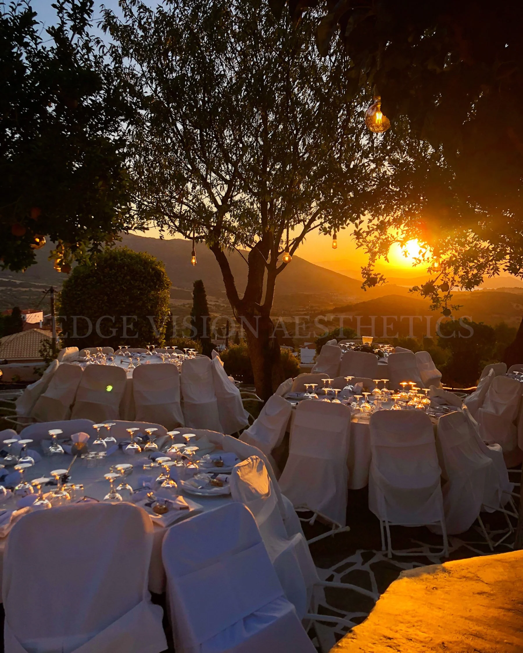 Outdoor event setup at sunset with rows of chairs covered in white cloth, a tree with hanging string lights, and a scenic landscape with hills in the background.
