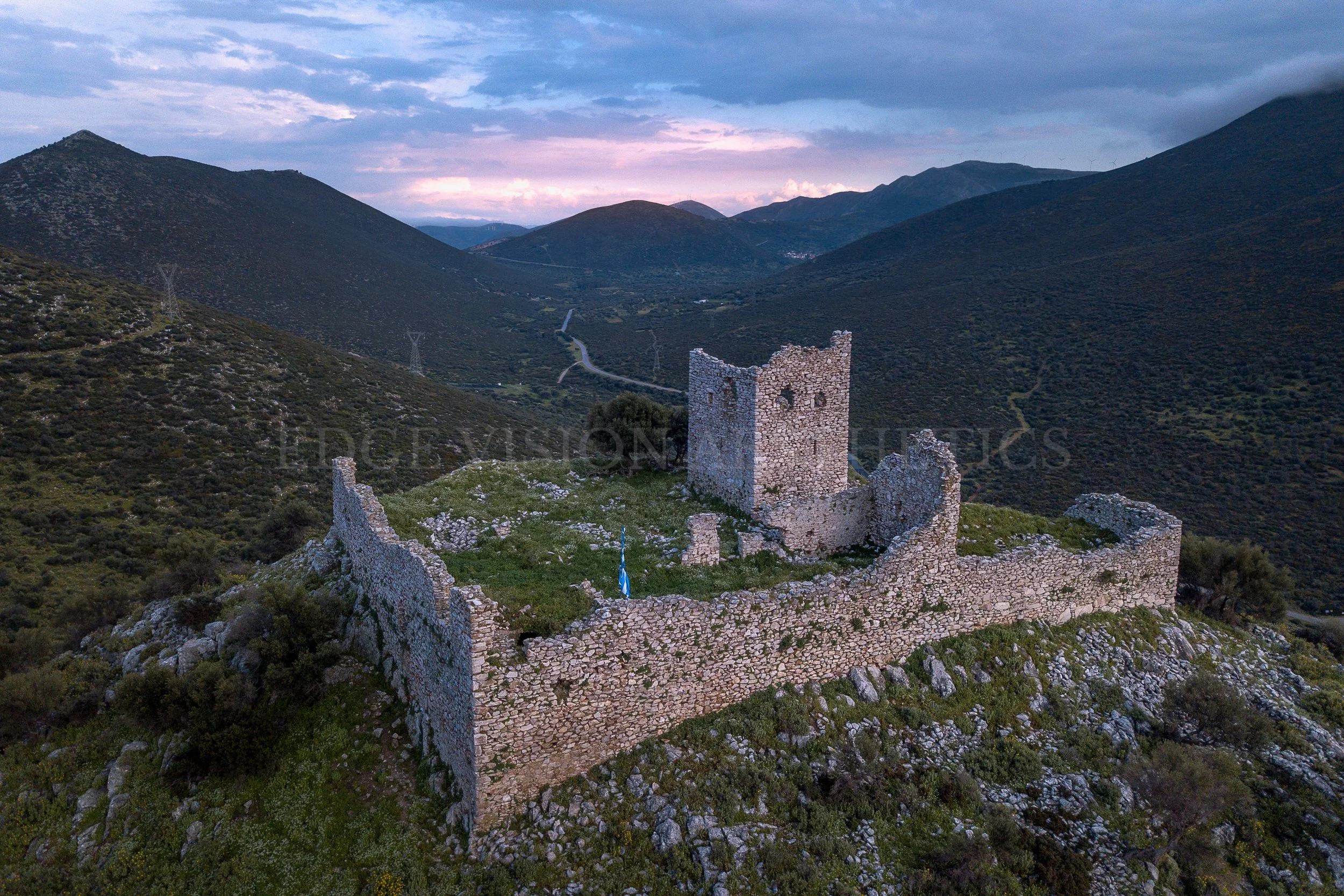 Ancient stone ruins of a castle or fortress on a mountain slope with hills and a cloudy sky in the background.