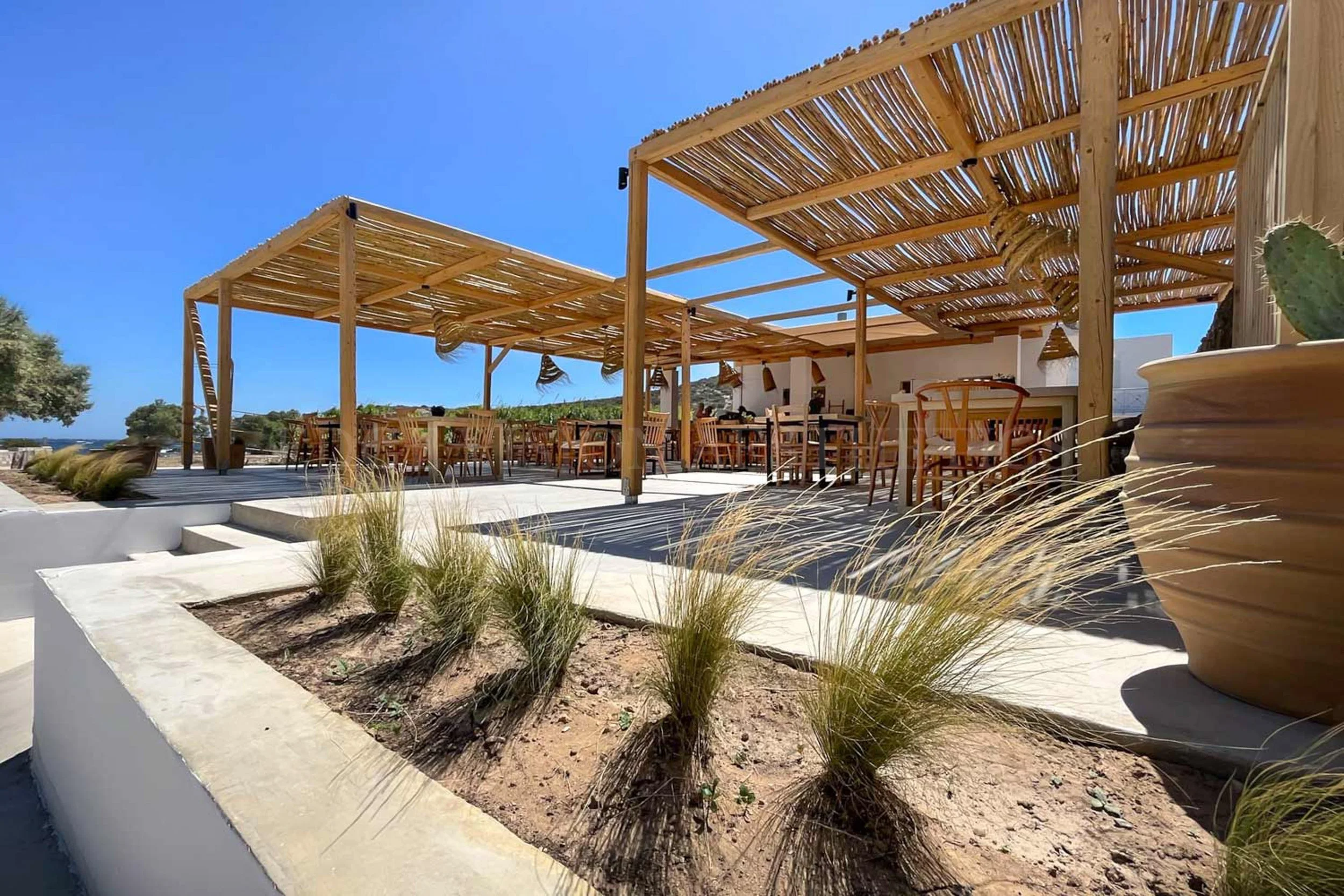 Outdoor patio with wooden pergola, seating area with chairs, potted cactus, and desert landscaping under a clear blue sky.