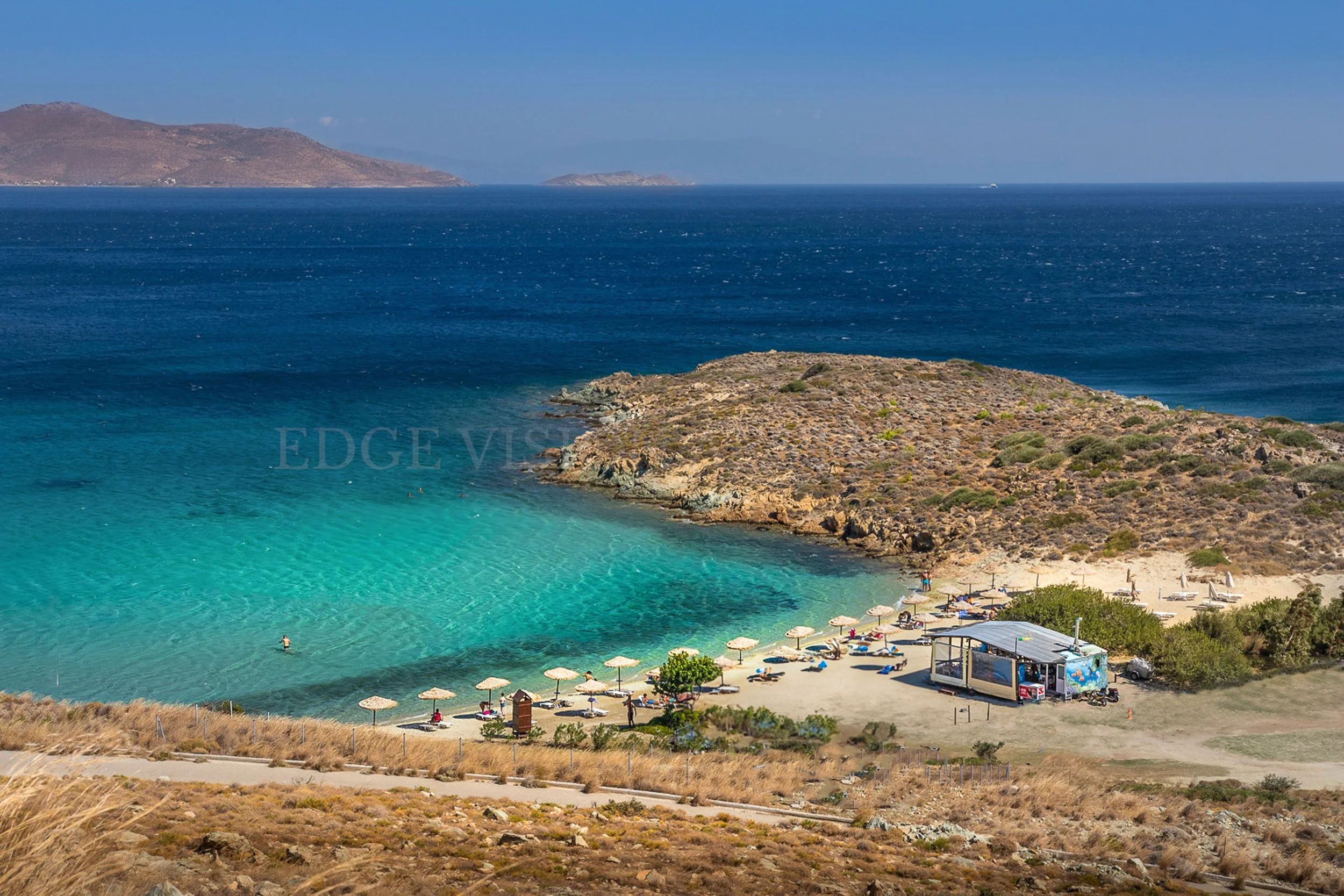 A rocky coastline with a sandy beach featuring umbrellas and lounge chairs, a small kiosk or snack stand, and a few people enjoying the clear turquoise water and sunny weather, with hills and islands in the background.