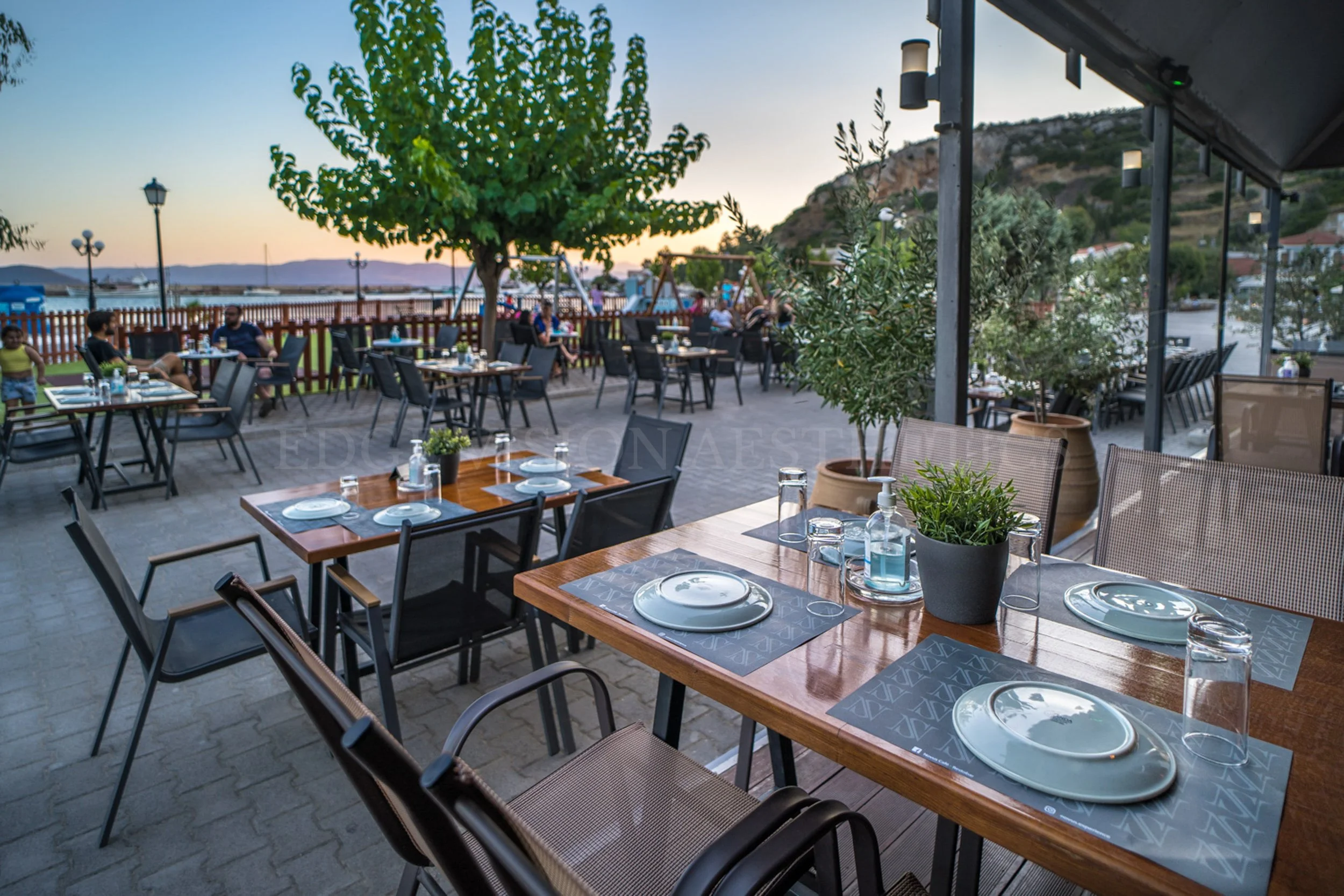 An outdoor dining area with tables set with plates, glasses, and potted plants, overlooking a waterfront with people sitting on a grassy area, trees, a fence, and distant mountains at sunset.