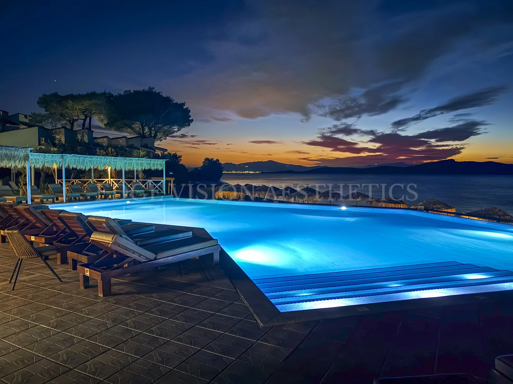 A nighttime view of a lit swimming pool with surrounding lounge chairs, overlooking a body of water and a sunset sky with clouds over mountains.