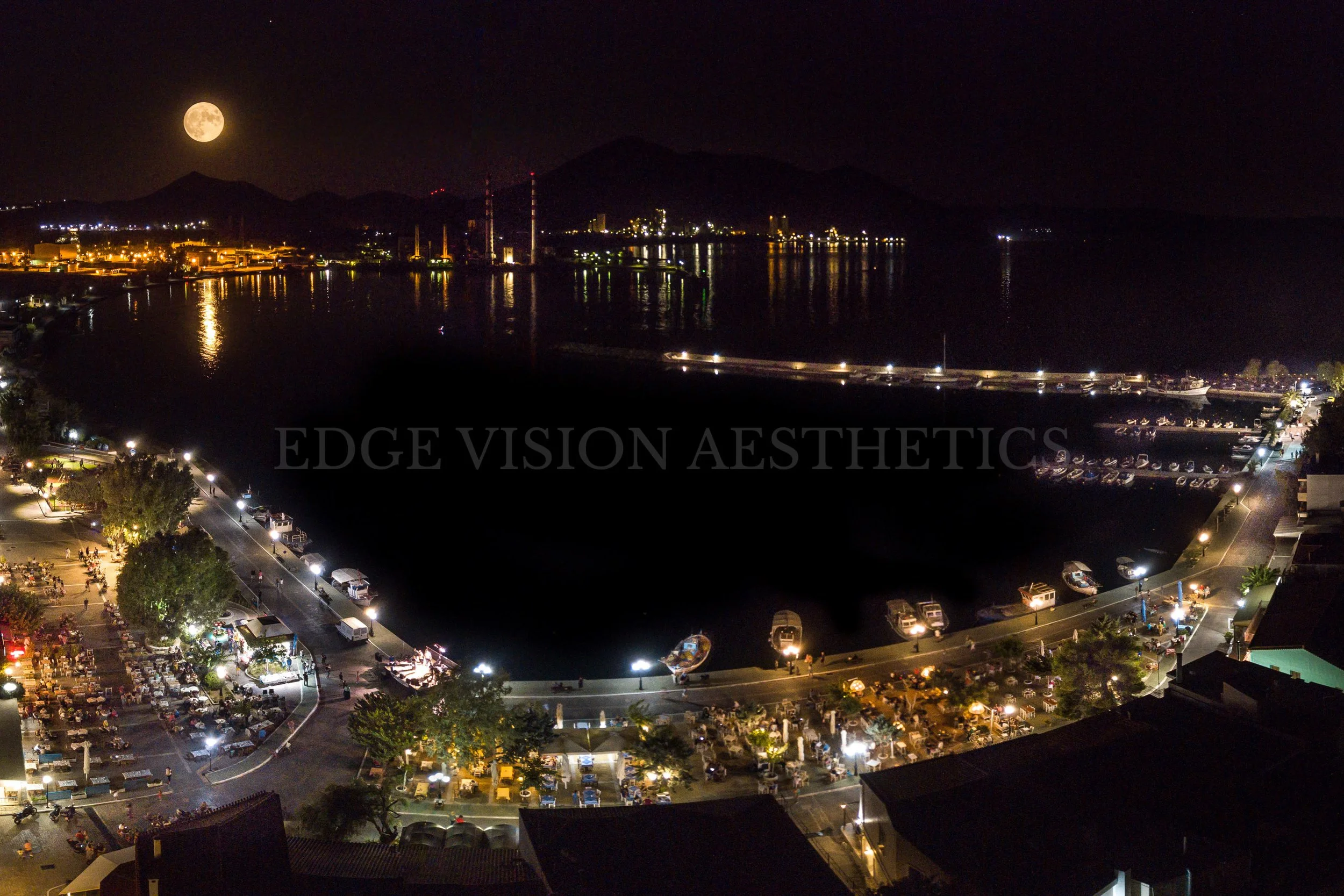 Night view of a harbor with boats and a parking lot filled with cars, illuminated by streetlights, with mountains and a full moon in the background.