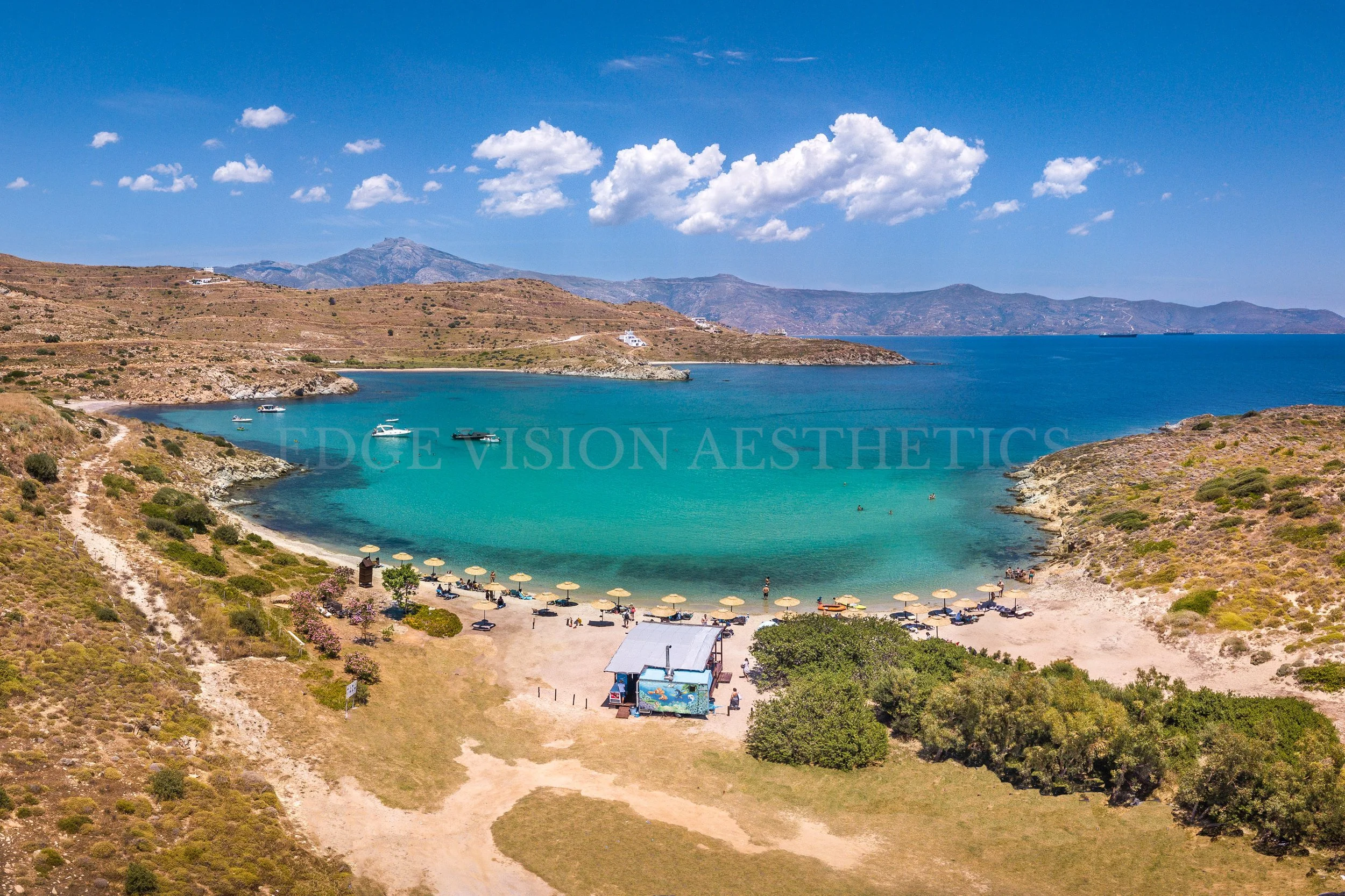 A scenic coastal landscape featuring a bay with turquoise waters, surrounded by arid hills and mountains in the background. There are boats on the water, a small beach with umbrellas, and a few park structures and trees along the shoreline under a br