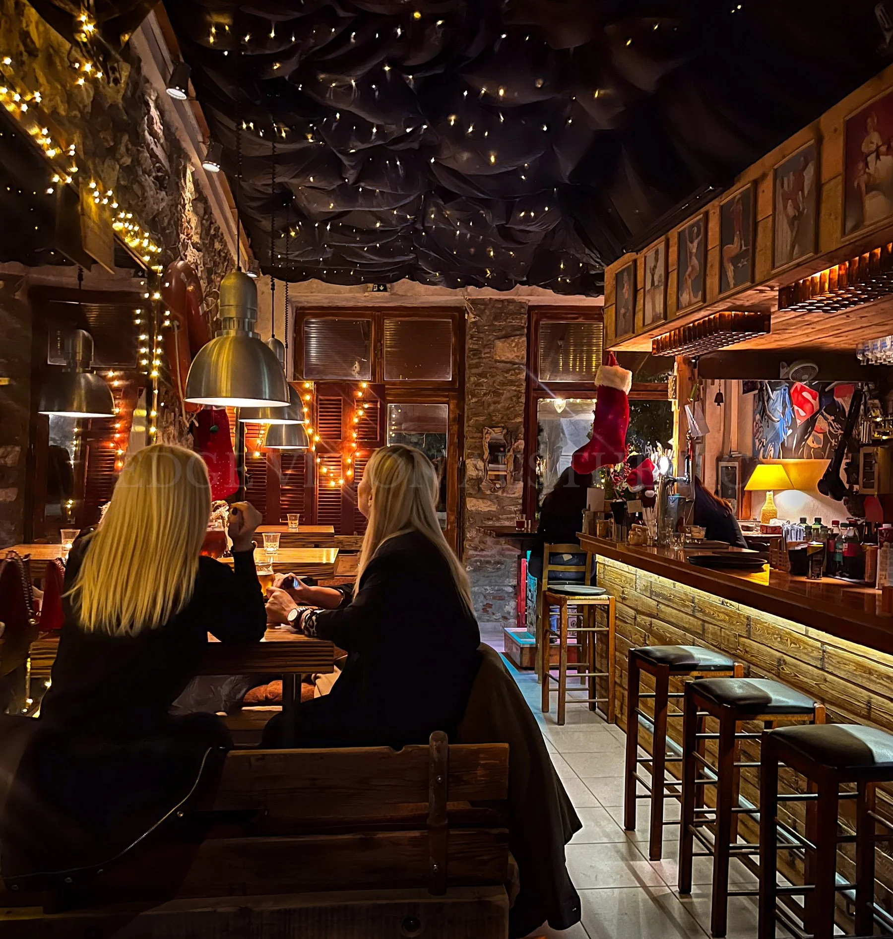 Interior of a cozy restaurant decorated with fairy lights, featuring wooden furniture, a stone wall, festive decorations including a Santa hat, and framed artwork on the wall.