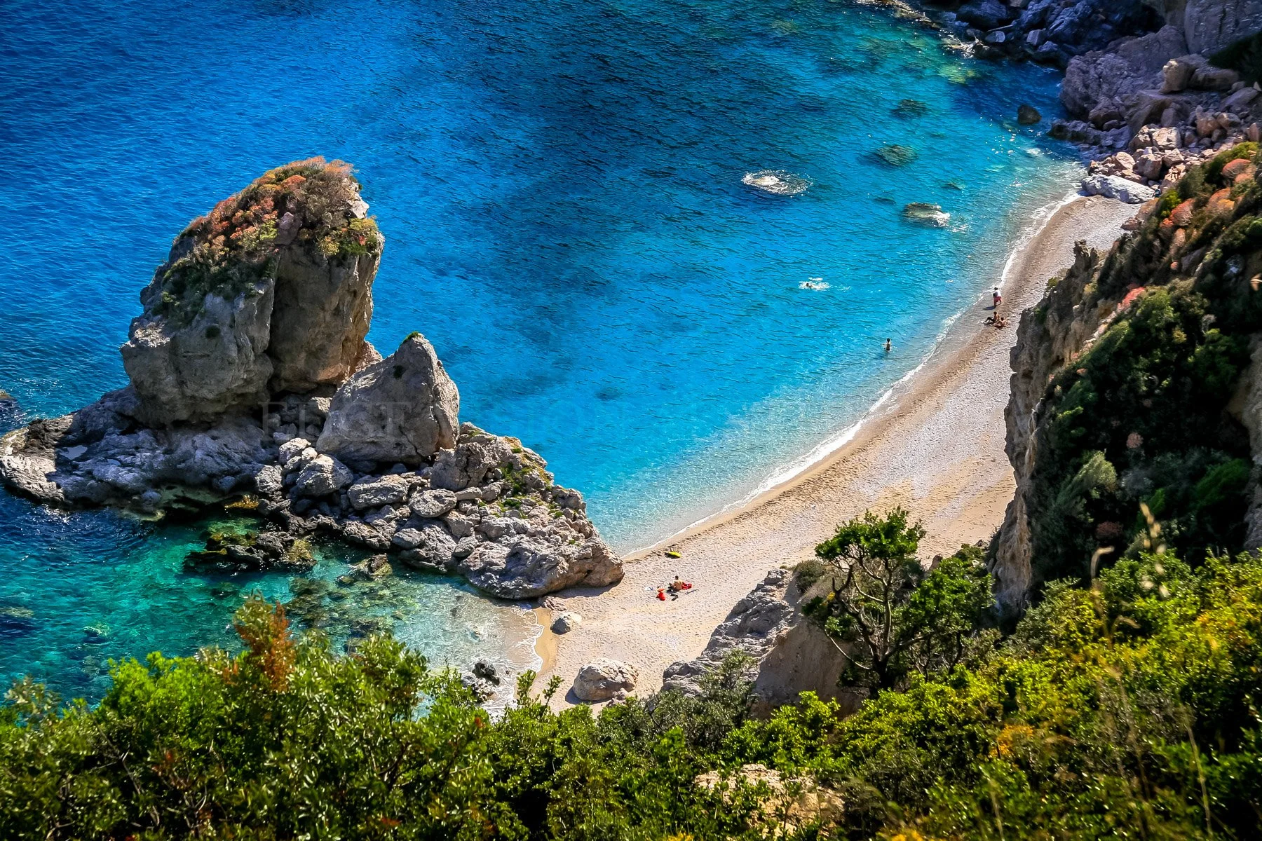 A secluded beach with clear turquoise water, large rocks, and green bushes on the cliffside.