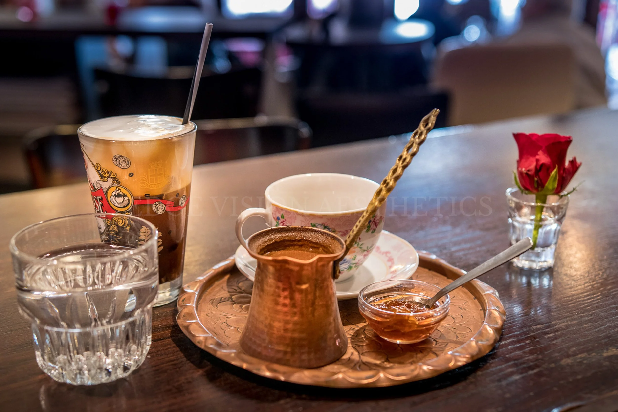 A table with various beverages and a small vase with a red flower. There is a glass of iced coffee, a porcelain cup with a floral pattern, a copper coffee pot, a small bowl of honey, and a small bowl of jam on a decorative tray. The background is blu