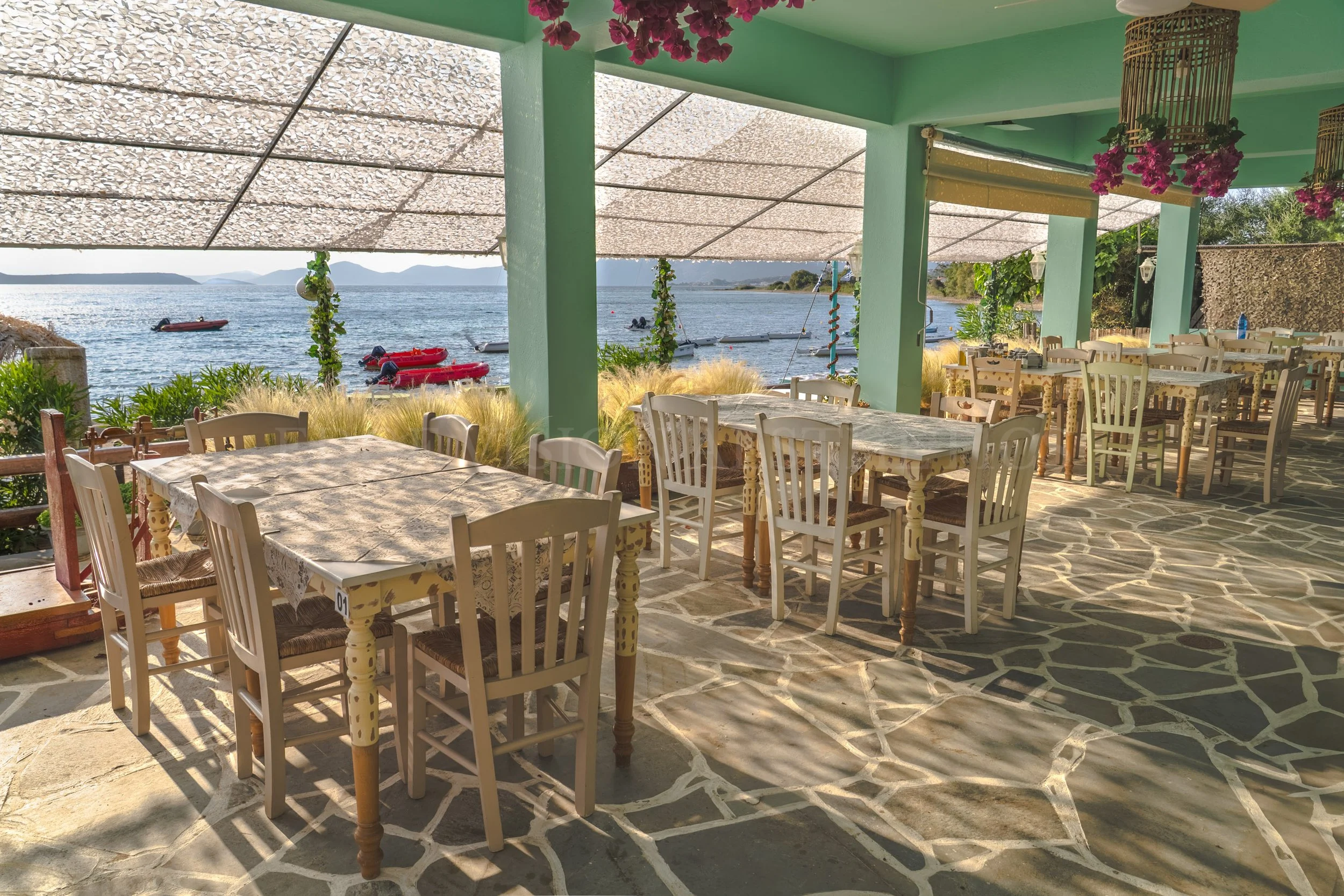 Outdoor restaurant seating area with tables and chairs overlooking the water, with boats and mountains in the distance, decorated with pink flowers and hanging lamps.