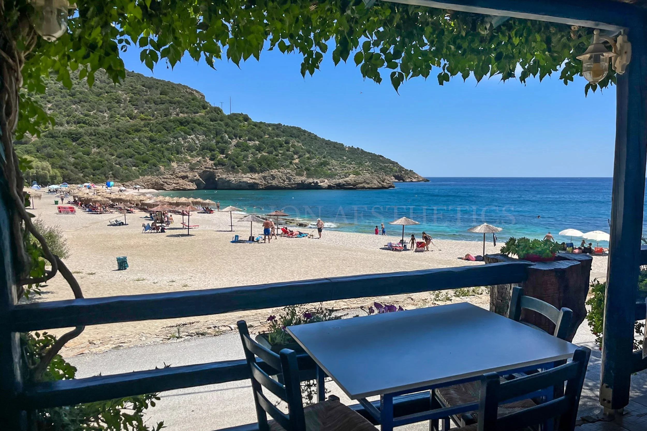 View from a shaded patio overlooking a sandy beach with lounge chairs and umbrellas, a calm sea, and a hill covered with green bushes and trees under a clear blue sky.