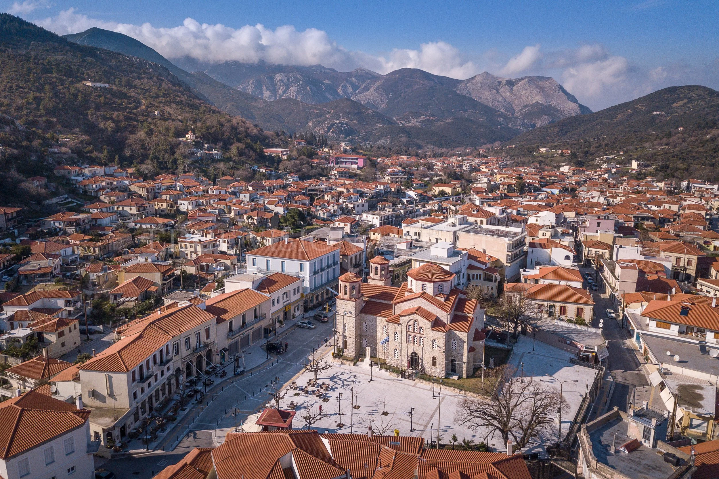 Aerial view of a town with red-tiled roofs, surrounded by mountains with some trees and buildings, under a partly cloudy sky.