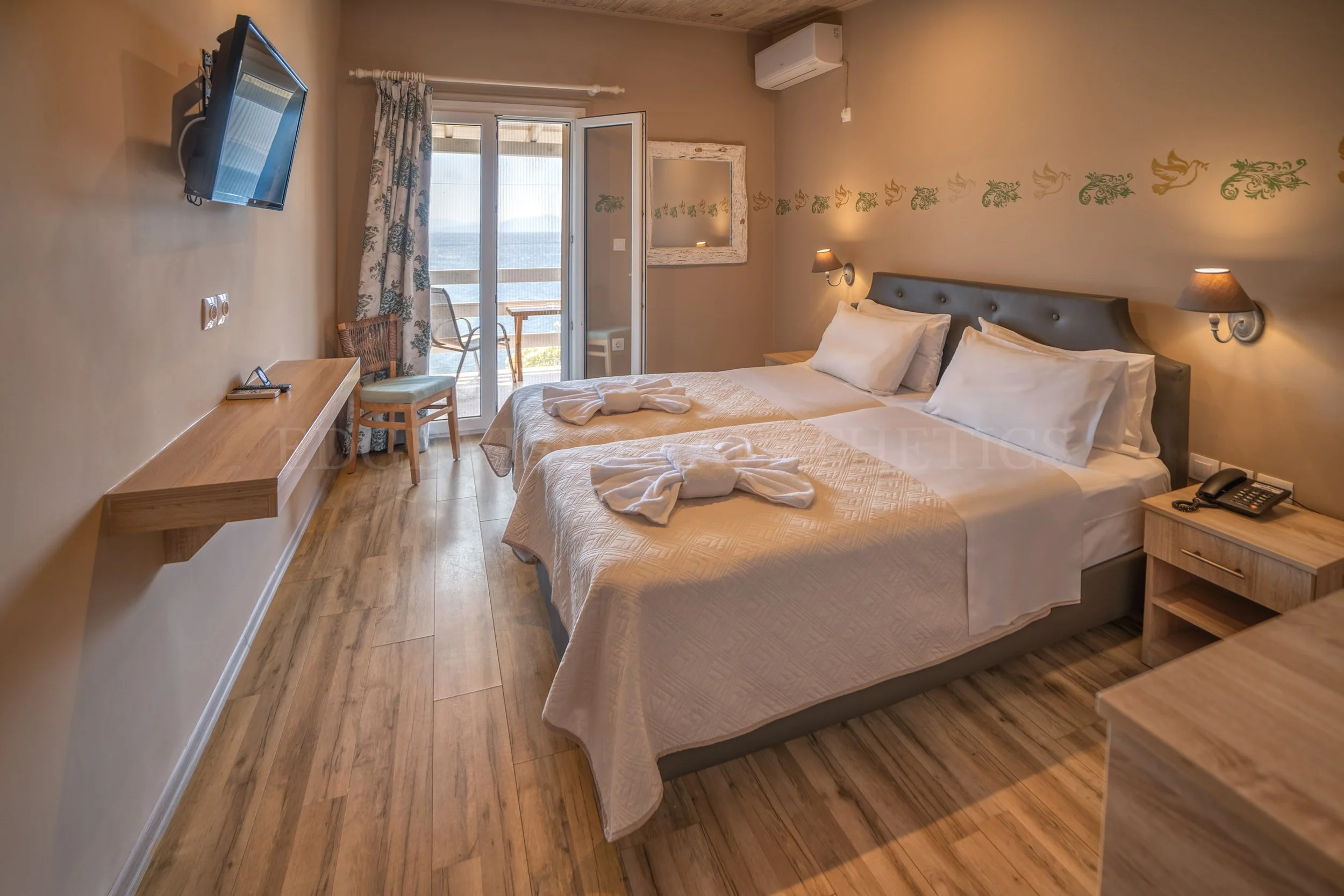 A hotel room with two single beds, wooden nightstand, wall-mounted lamps, a flat-screen TV, a sliding glass door leading to a balcony, and mountain view outside.