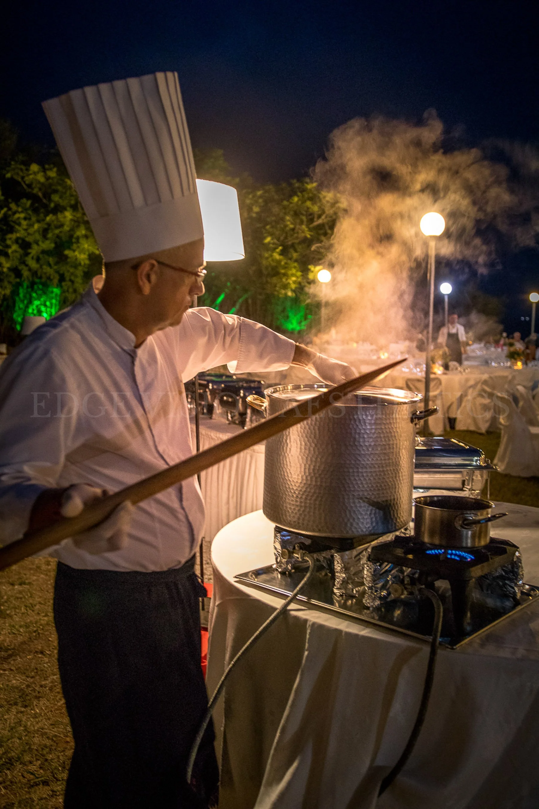 A chef stirring a large pot on an outdoor stove at night, with steam rising, in a decorated outdoor event setting.
