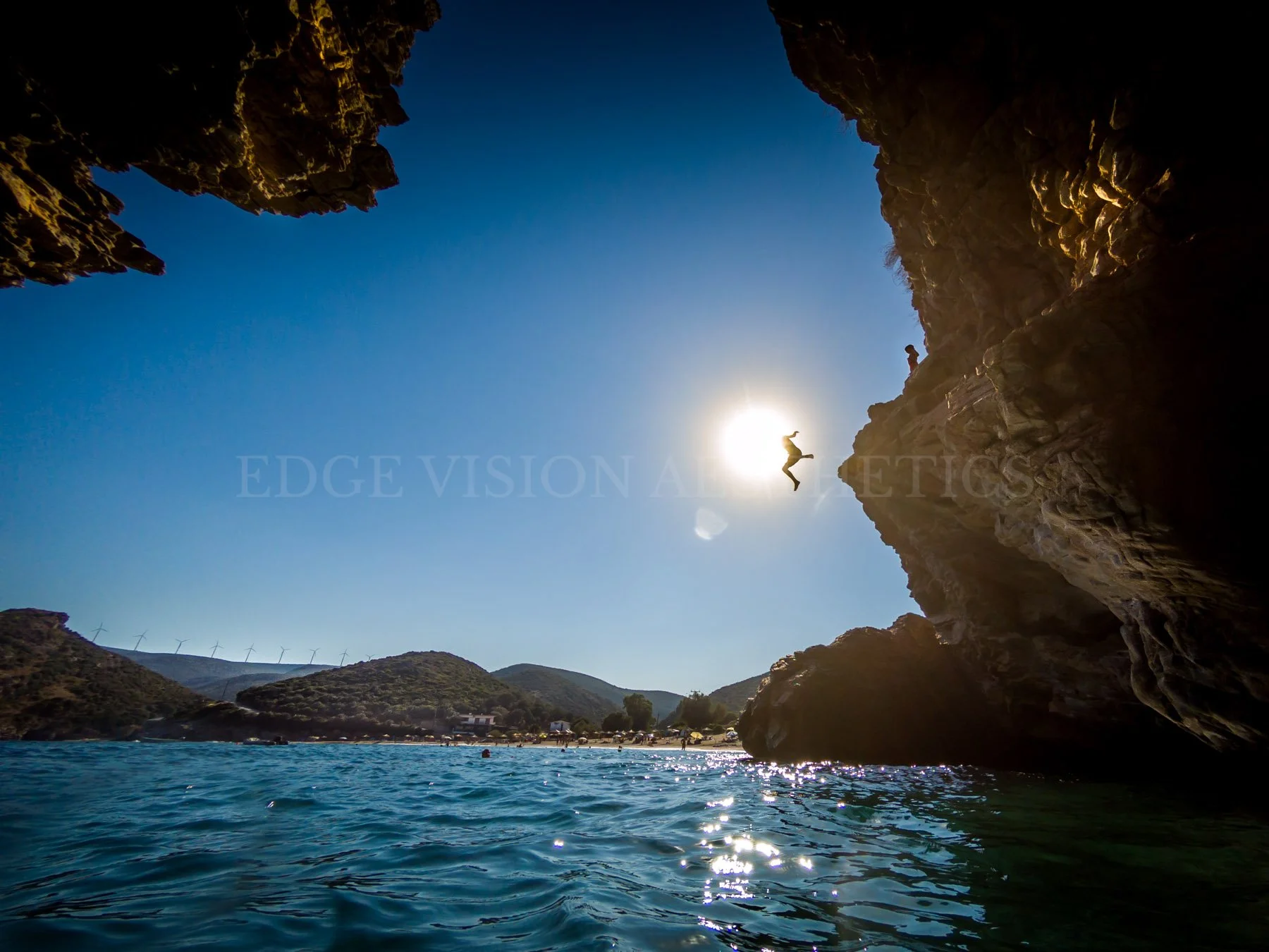 View from inside a rocky cave looking out over a body of water with hills and wind turbines in the distance, person jumping off the cave ledge against the bright sun.