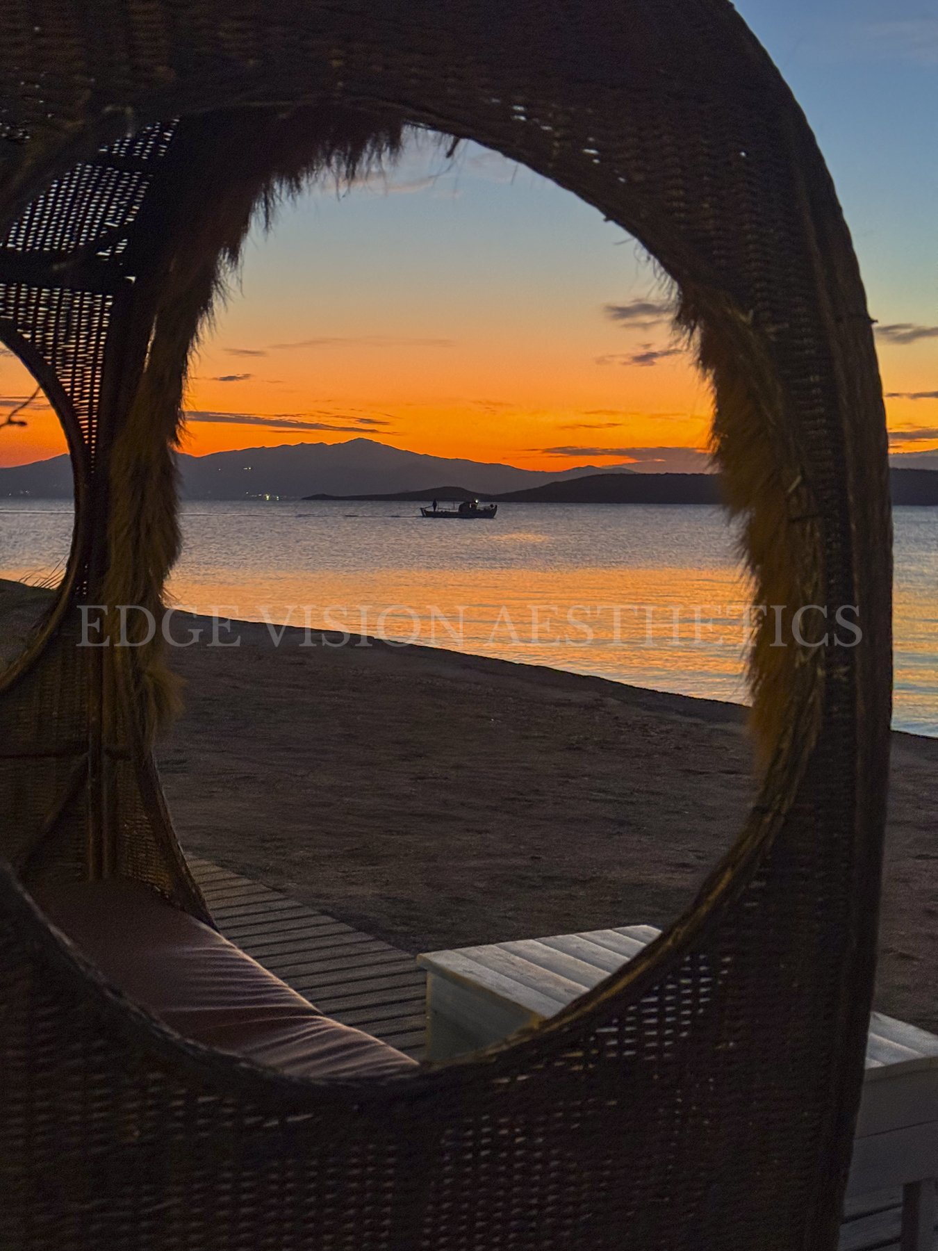 Sunset over a lake with mountains in the background, seen through a rattan mirror frame on a deck with a small bench.