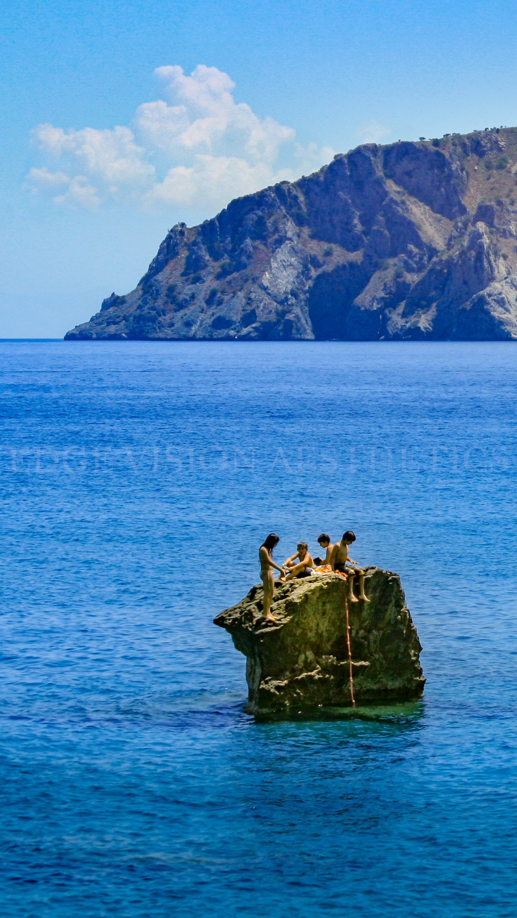 Four children sitting on a large rock in the ocean, with a mountainous island and partly cloudy sky in the background.