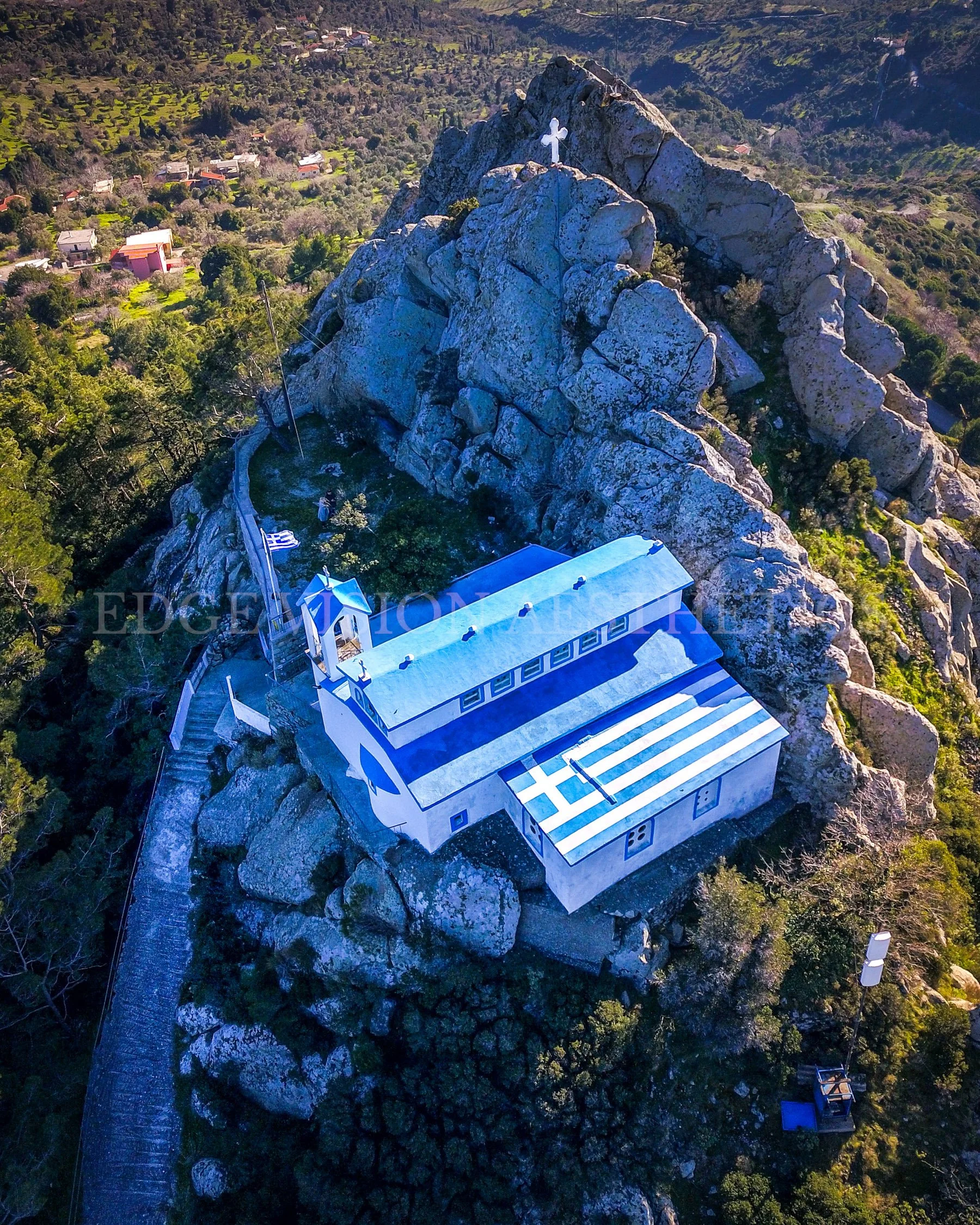 An aerial view of a rocky mountain with a white and blue chapel built on the slope. The chapel has a cross on top and a staircase leading to the entrance. There is a Greek flag at the base of the mountain. Surrounding the mountain are trees, houses, 