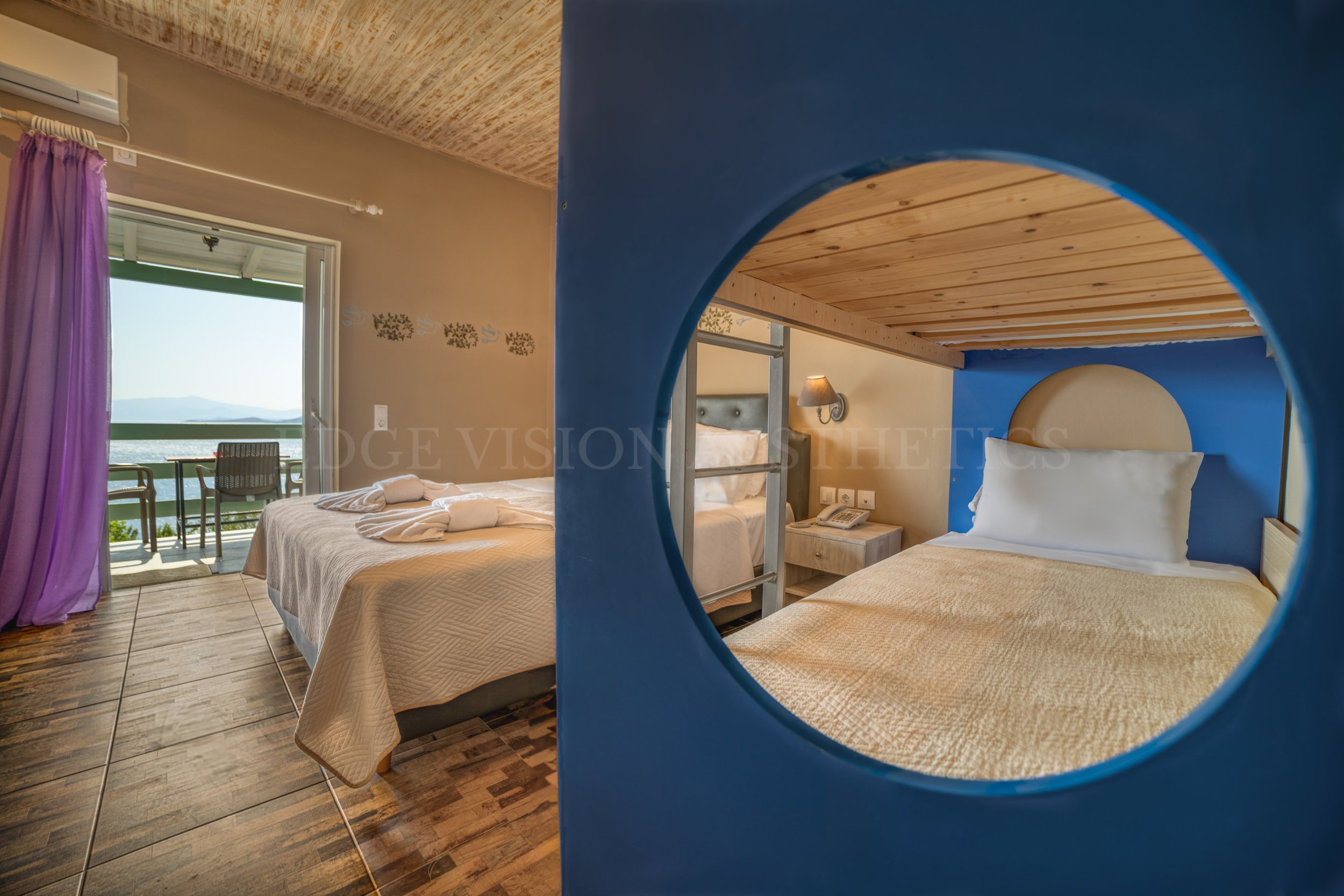 View of a hotel room with two beds, a balcony with chairs, and a scenic mountain landscape visible through the window. The photo is taken through a circular blue window.