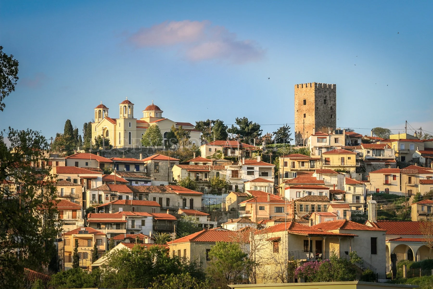 View of a hillside town with beige and white houses topped with red tile roofs, a yellow church with three bell towers, and a stone tower on a hill under a blue sky with scattered clouds.