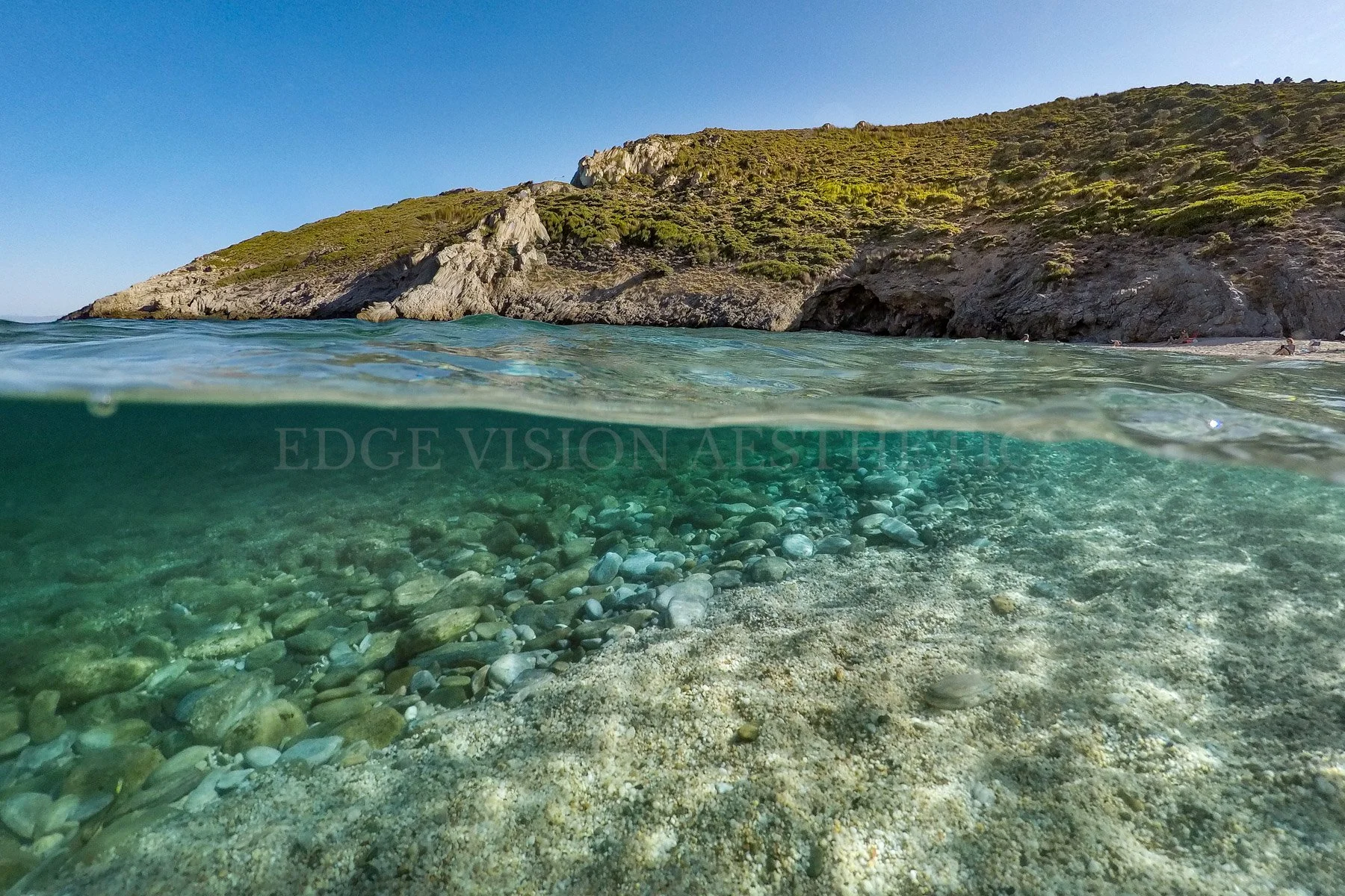 A scenic view of a beach with clear water, pebbles, a rocky hillside, and a blue sky.
