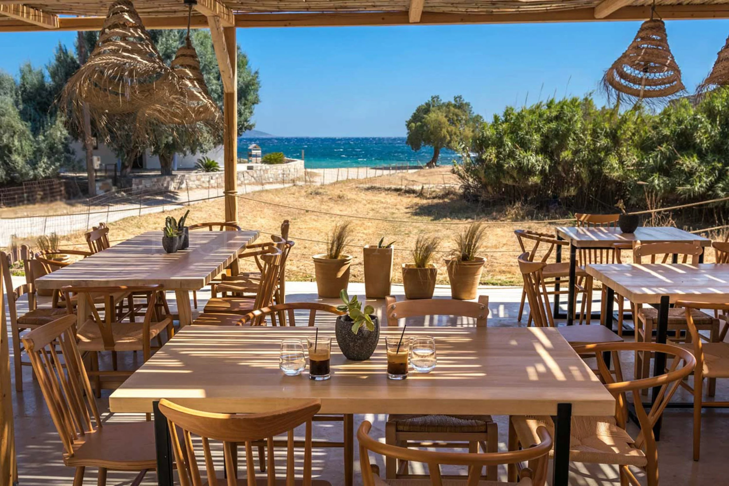 Wooden outdoor dining area overlooking sandy beach and blue ocean, with trees and shrubs in the background, under a wooden pergola with woven lampshades.
