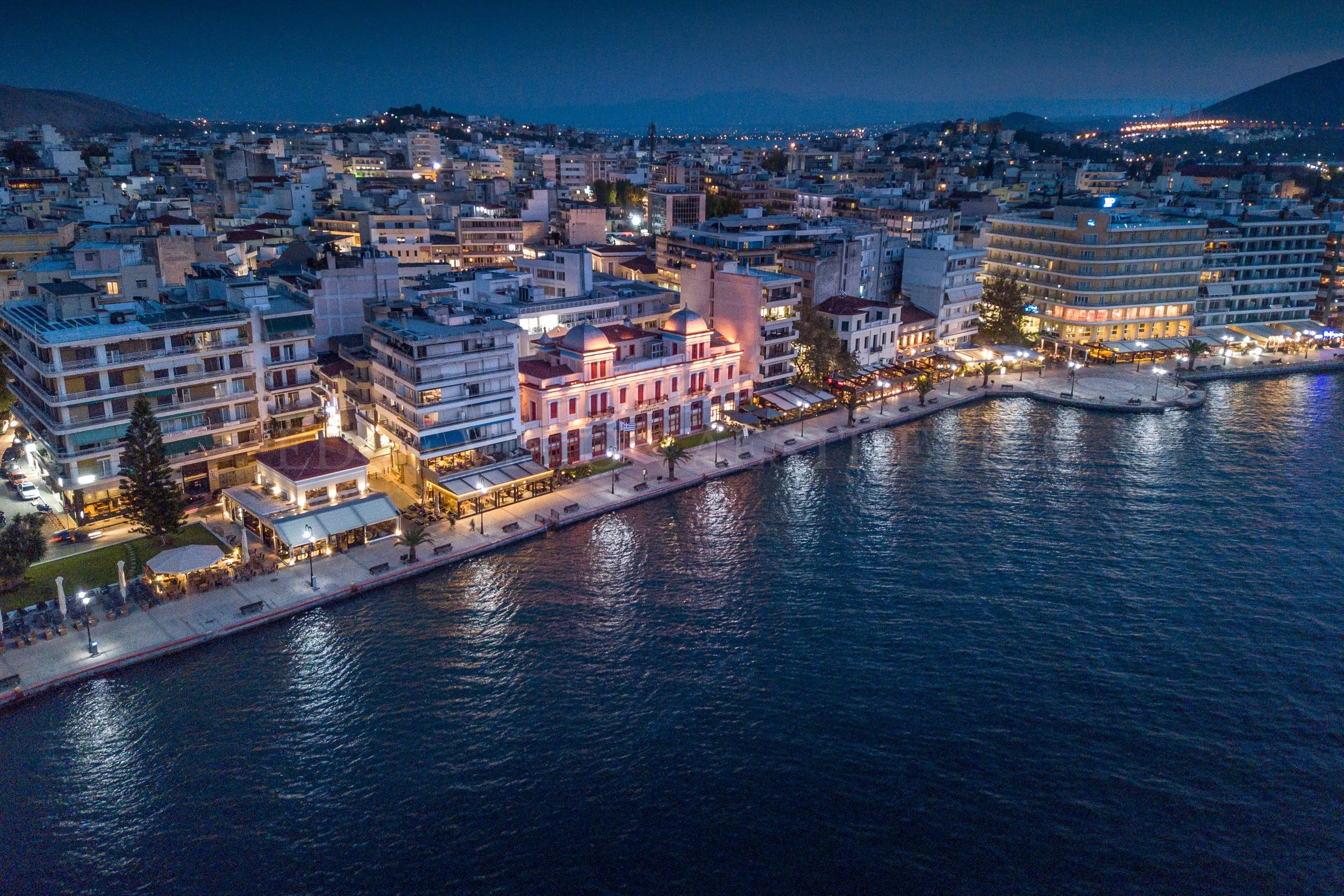 Aerial view of a city by the water at night, showing lit buildings along a waterfront promenade.