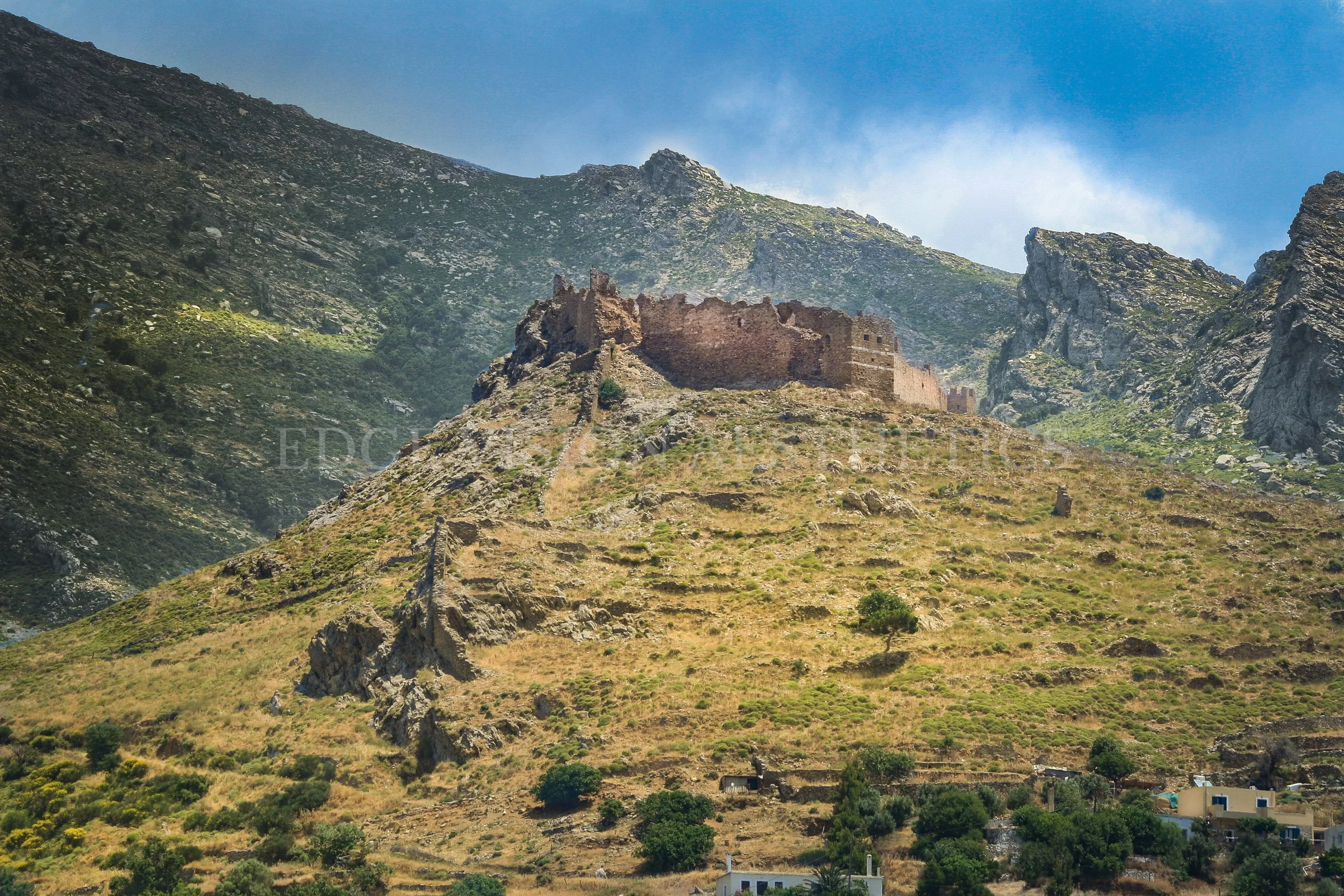 Ancient ruins perched on a hillside within a mountainous landscape under a blue sky.